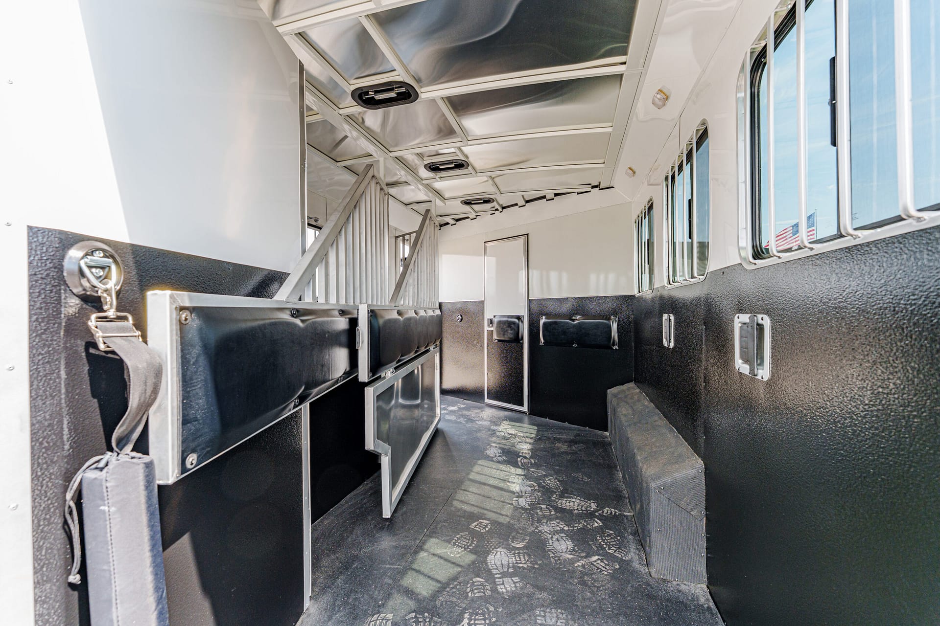 Inside a horse trailer with black padded stalls, metal dividers, and windows along the right wall