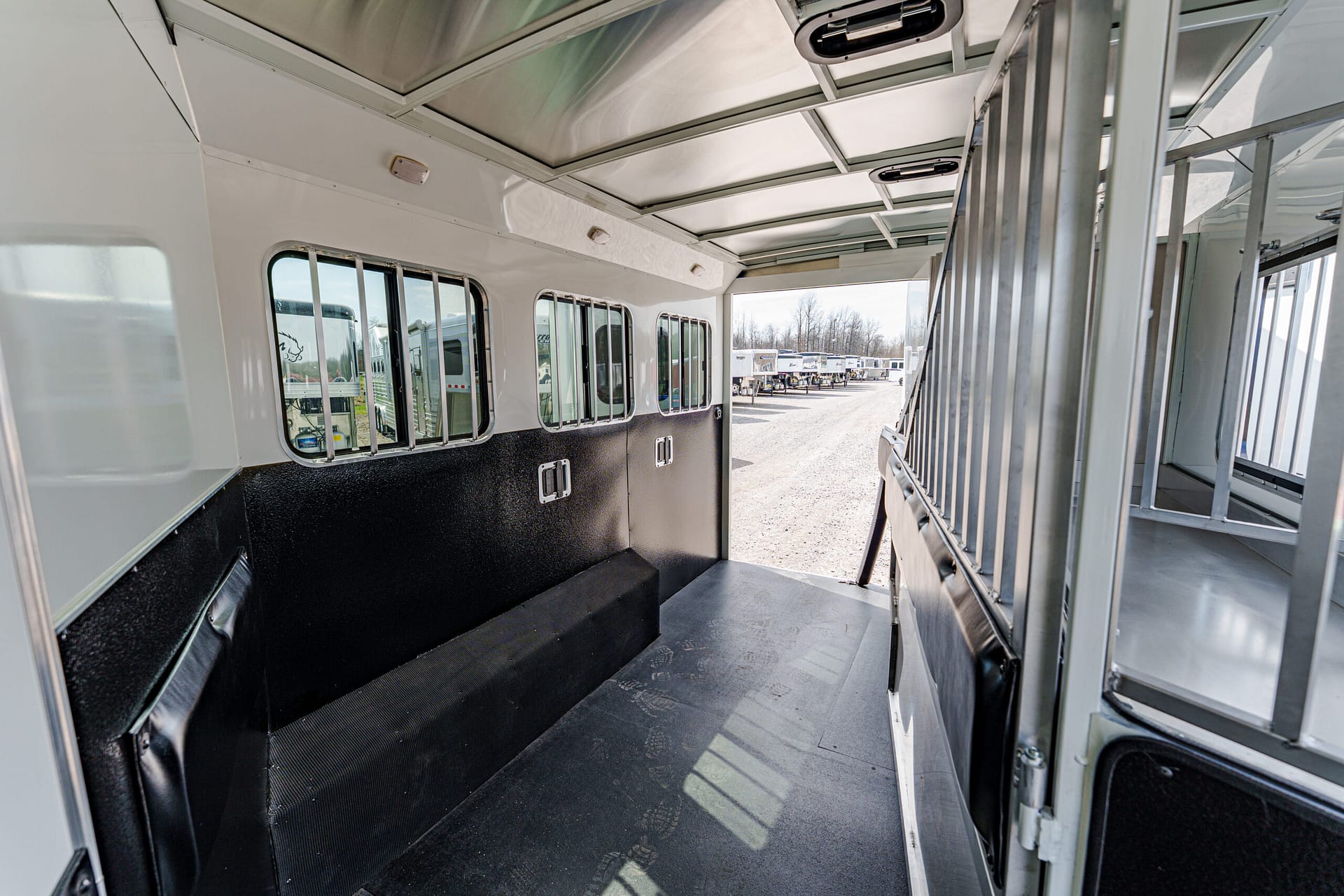 Interior of a white horse trailer with padded black walls, window bars along the left, and a wide open doorway to a gravel lot with parked trailers beyond.