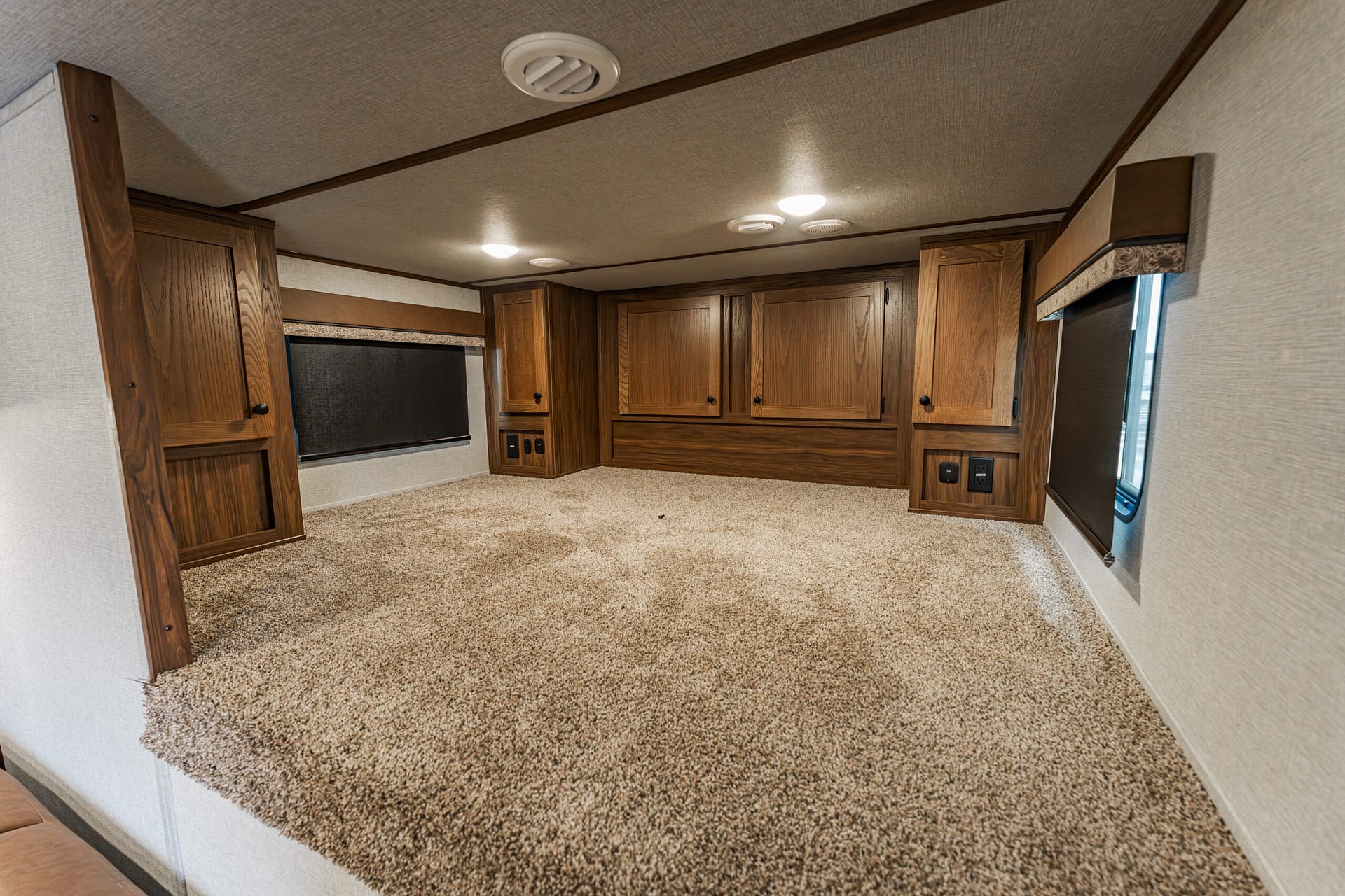 Rear RV bedroom with dark wood cabinets, beige carpet, and window shades along the back wall for storage and seating space.