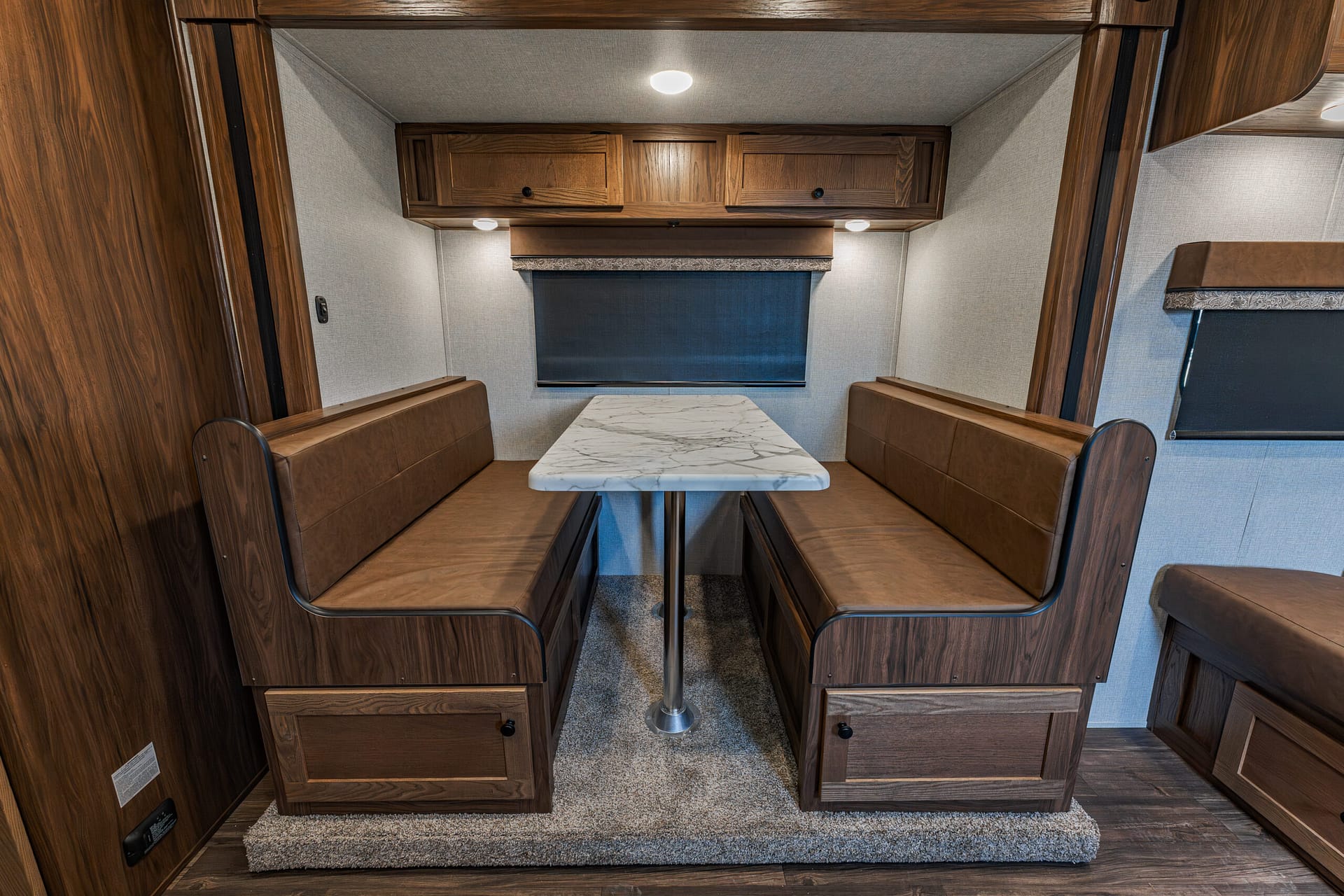 Booth-style dinette in a camper with a marble tabletop and brown leather benches facing each other under overhead wooden cabinets.