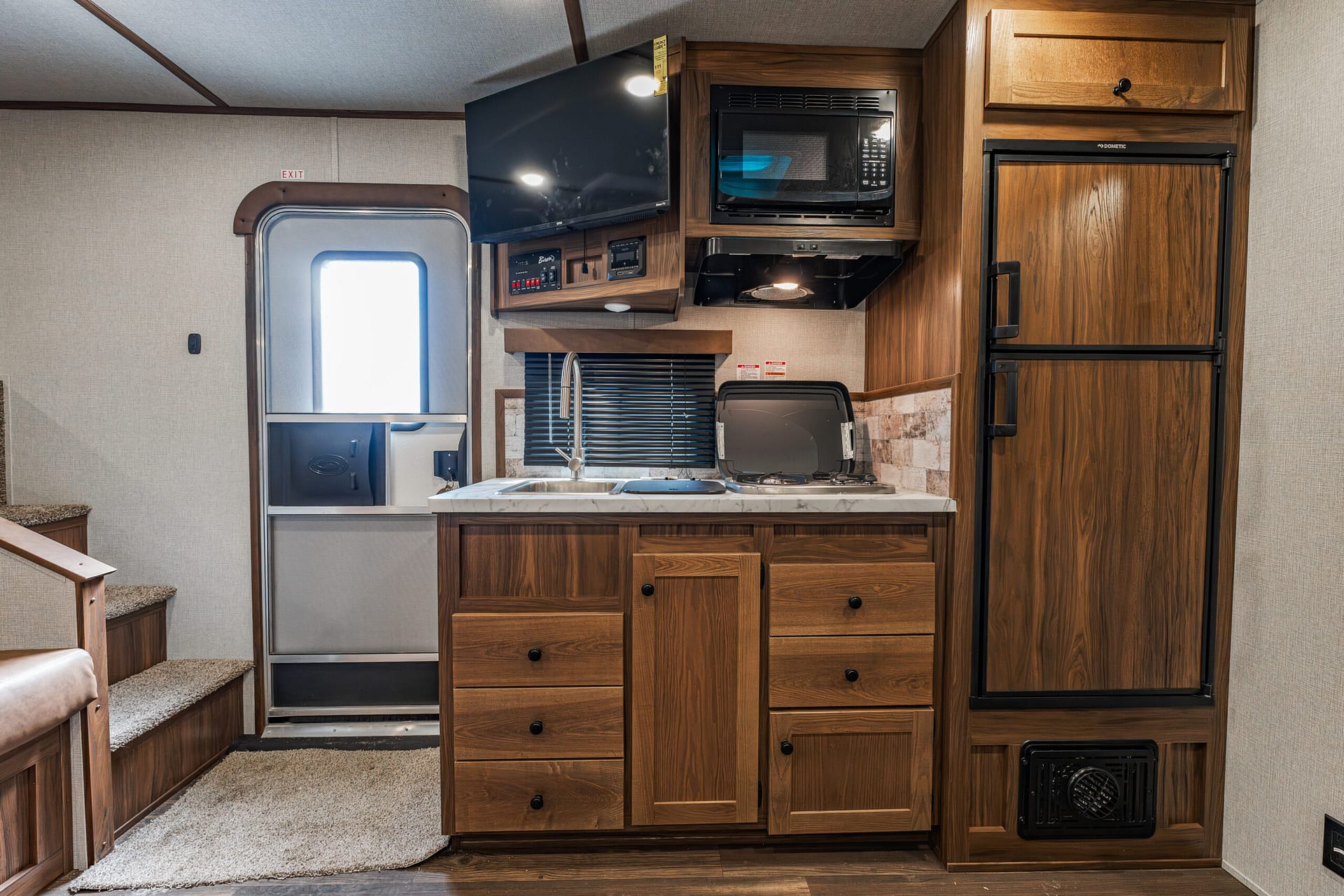 Wood-paneled RV kitchen with fridge, microwave, and sink along a marble counter top; doorway to exterior left.