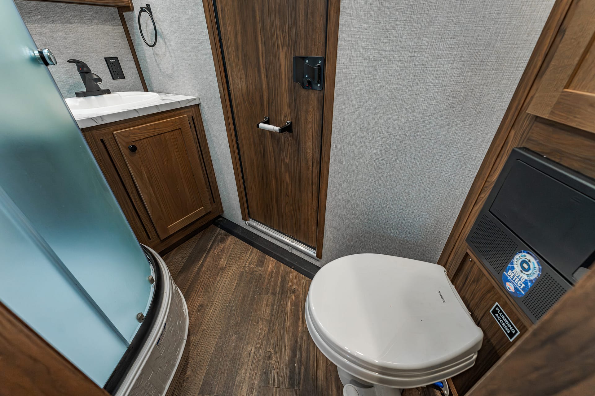 Compact RV bathroom with a wooden vanity, white sink, black faucet, frosted glass shower, and toilet nearby.