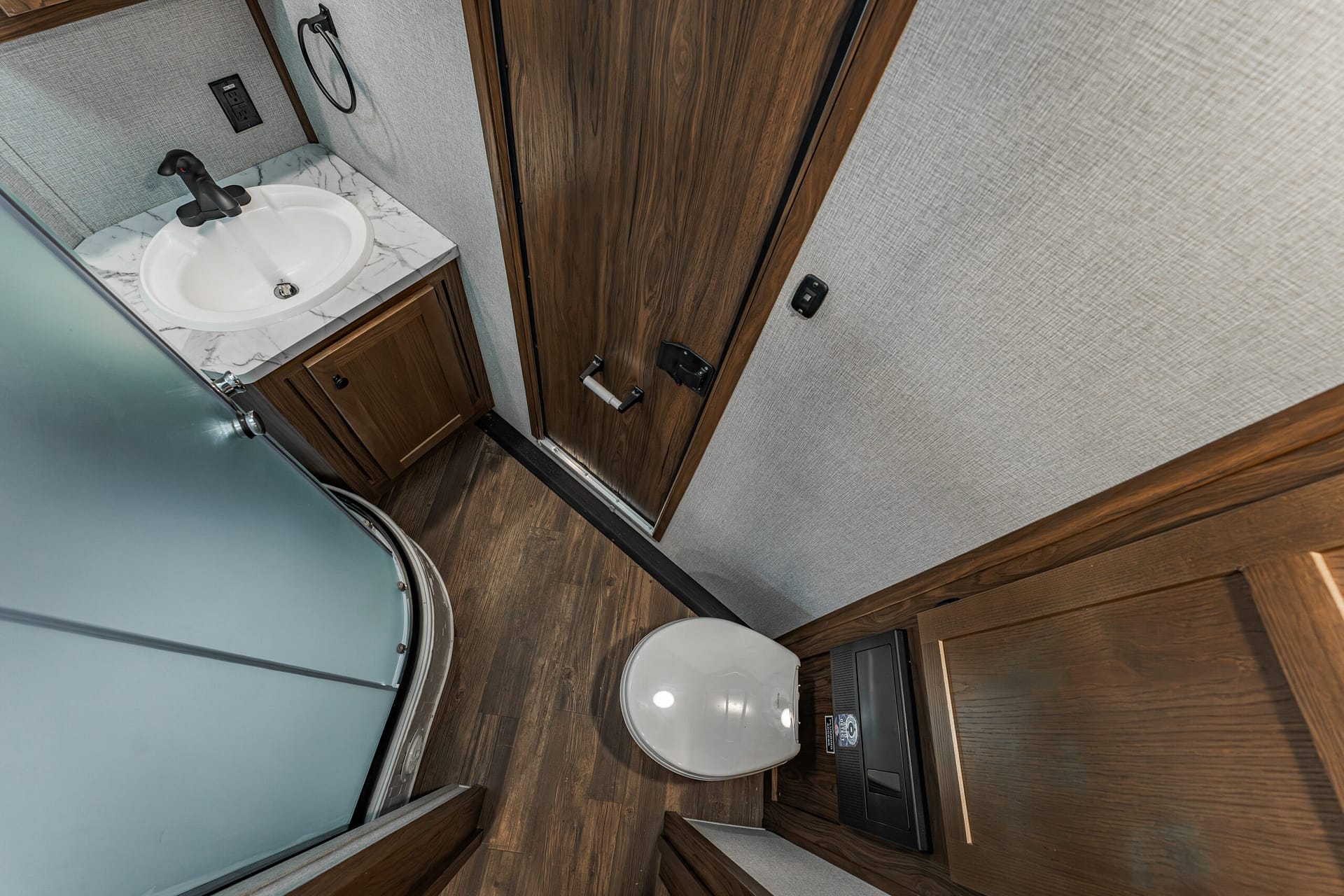 Compact RV bathroom with an oval sink, marble countertop, wooden vanity, and closed toilet next to a wooden door and glass shower enclosure.