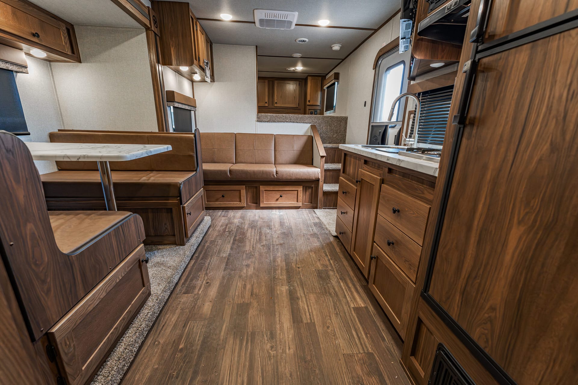 Interior of a wood-panelled RV with a brown dining booth, sofa, and kitchen along the right wall. Leather seating and cabinets throughout.