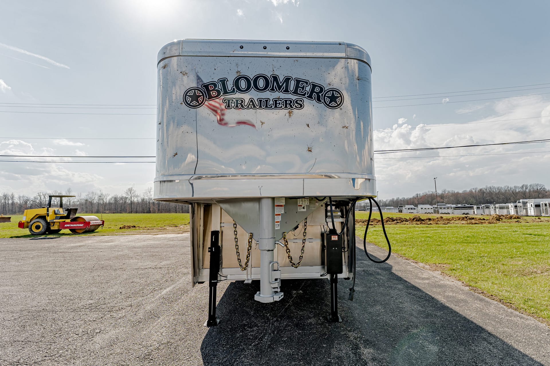 Shiny silver Bloomer Trailers gooseneck trailer on an asphalt lot with a yellow roller machine in the background and open fields beyond.