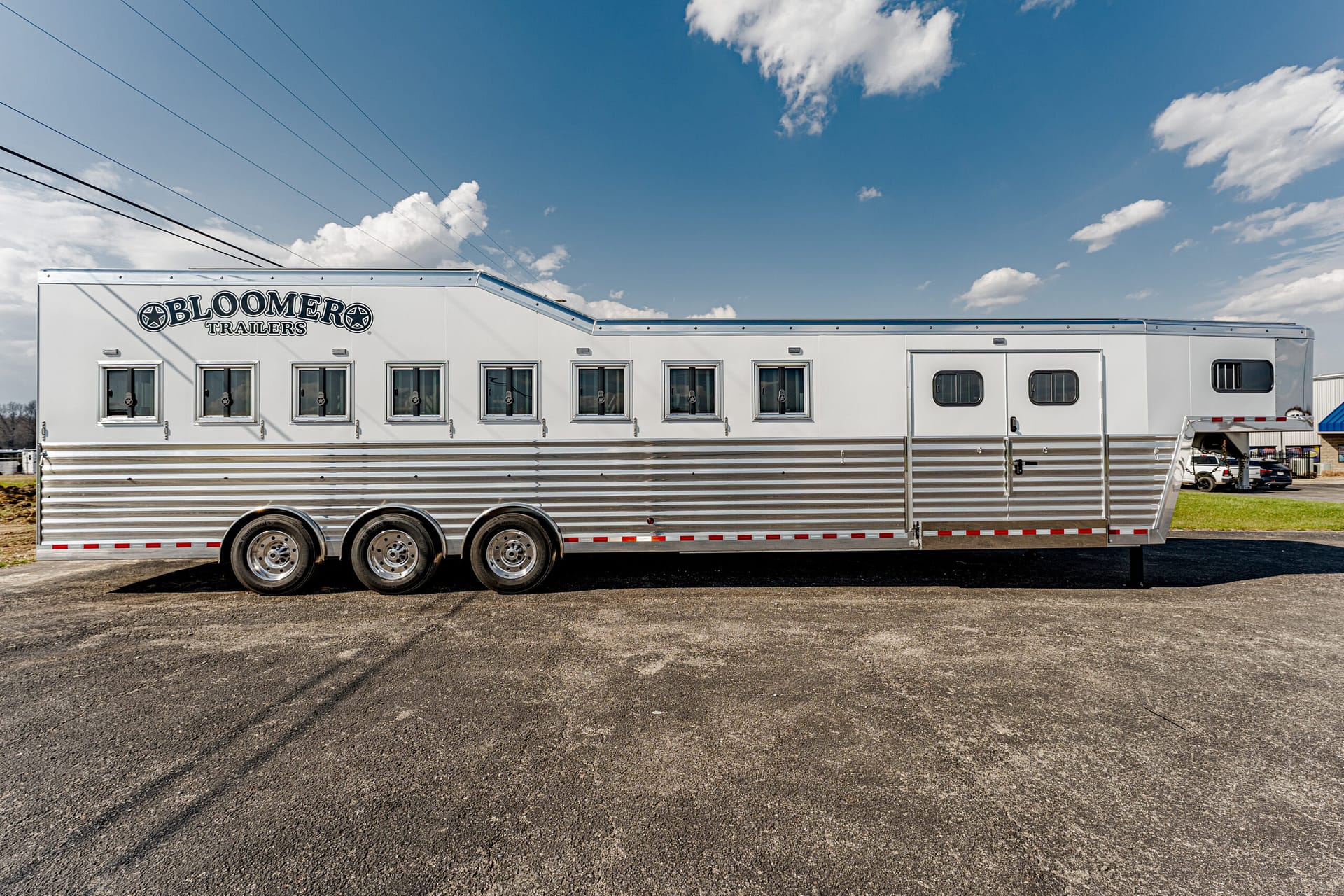 White Bloomer Trailers horse trailer with three axles and a row of windows, parked on asphalt under a blue sky with clouds.