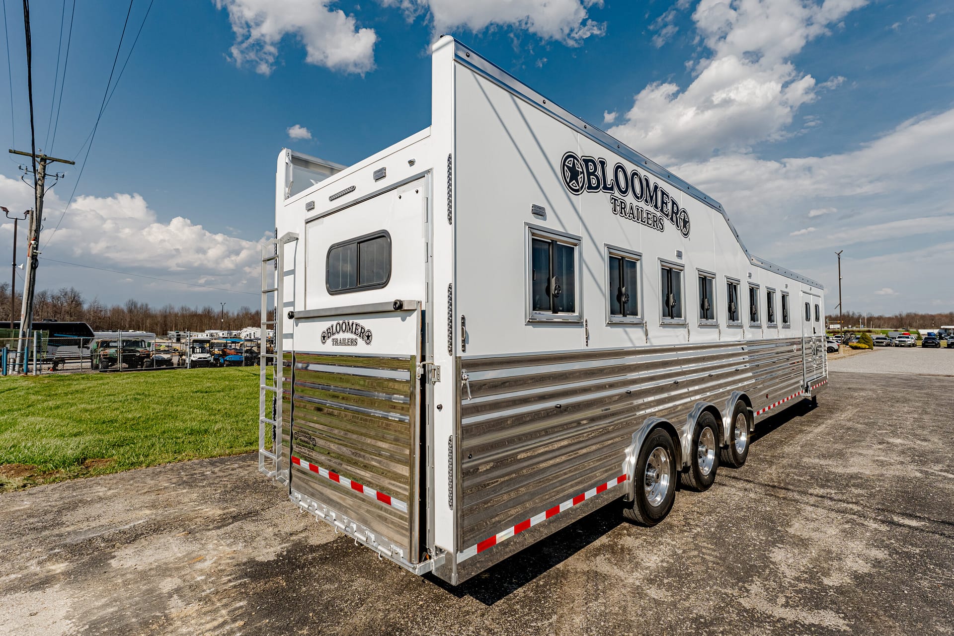 White Bloomer horse trailer with chrome lower siding parked on a paved lot under a blue sky with clouds; multiple windows along the side and a rear ramp gate visible.