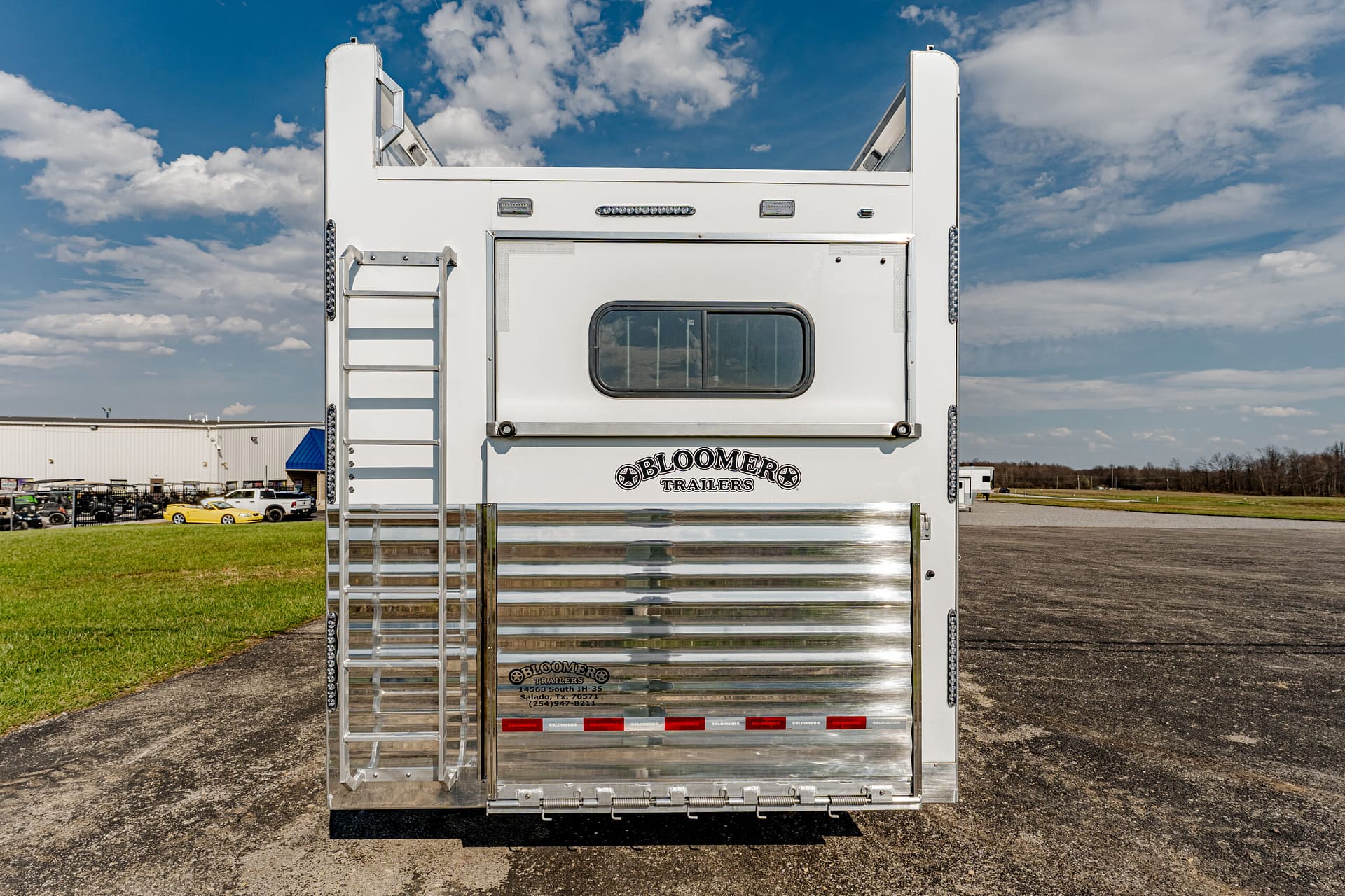 Back view of a white trailer with a metal ladder on the left, a small window, and chrome ribbed panel; 'Bloomers Trailers' logo visible on the back.