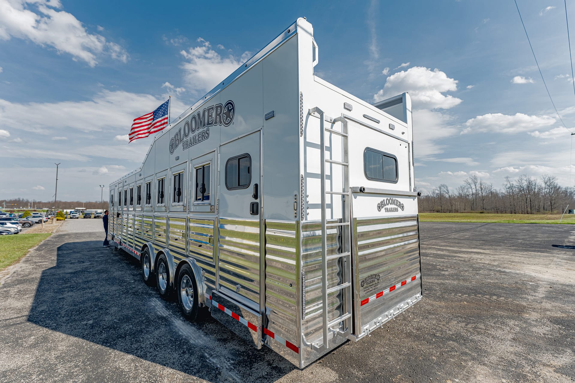 Rear three-quarter view of a tall aluminum Bloomers Trailers horse trailer with a ladder, parked on a lot under a blue sky with an American flag nearby.