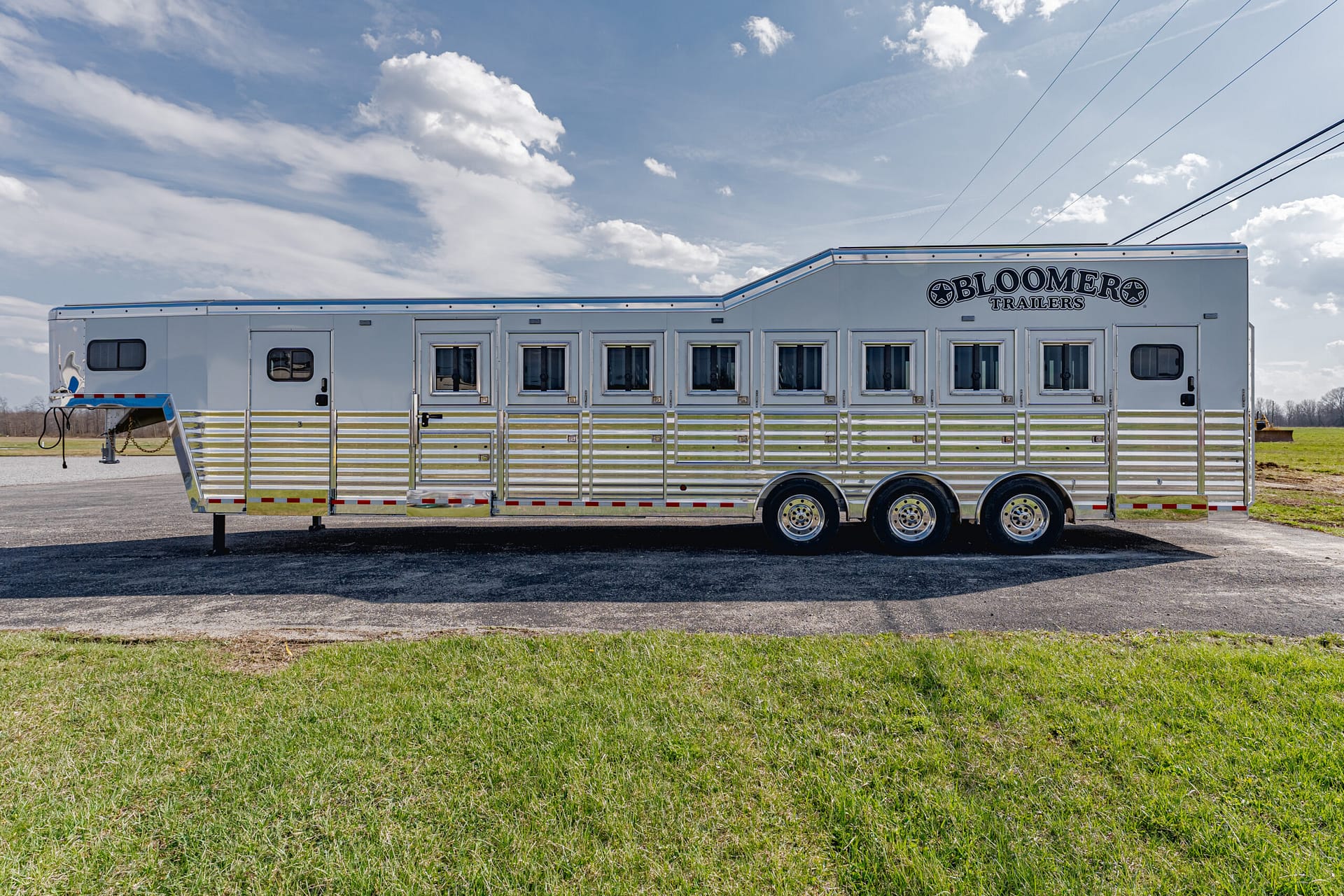 Silver aluminum horse trailer with multiple stalls and a row of windows, parked on a paved lot under a blue sky; Bloomers Trailers logo near the front.