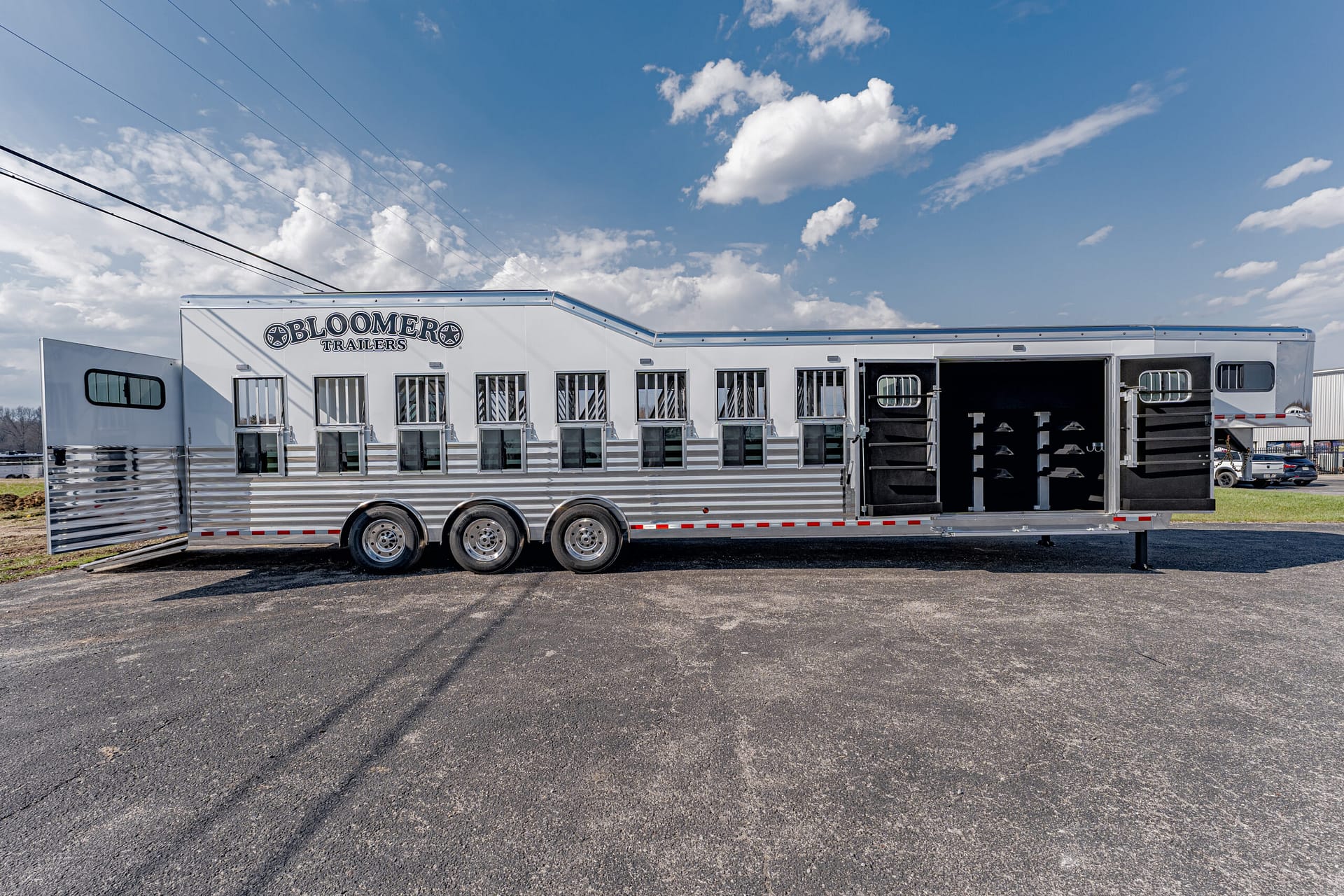 Long white and silver Bloomer horse trailer with multiple stalls and open rear doors, parked on asphalt under a blue sky with clouds.