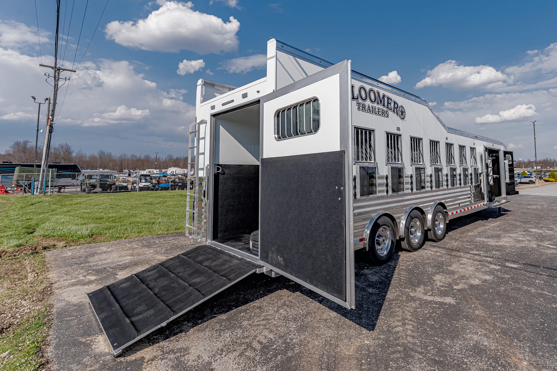 White Loomer horse trailer with rear ramp lowered for loading on a paved lot; interior stalls visible in black lining.