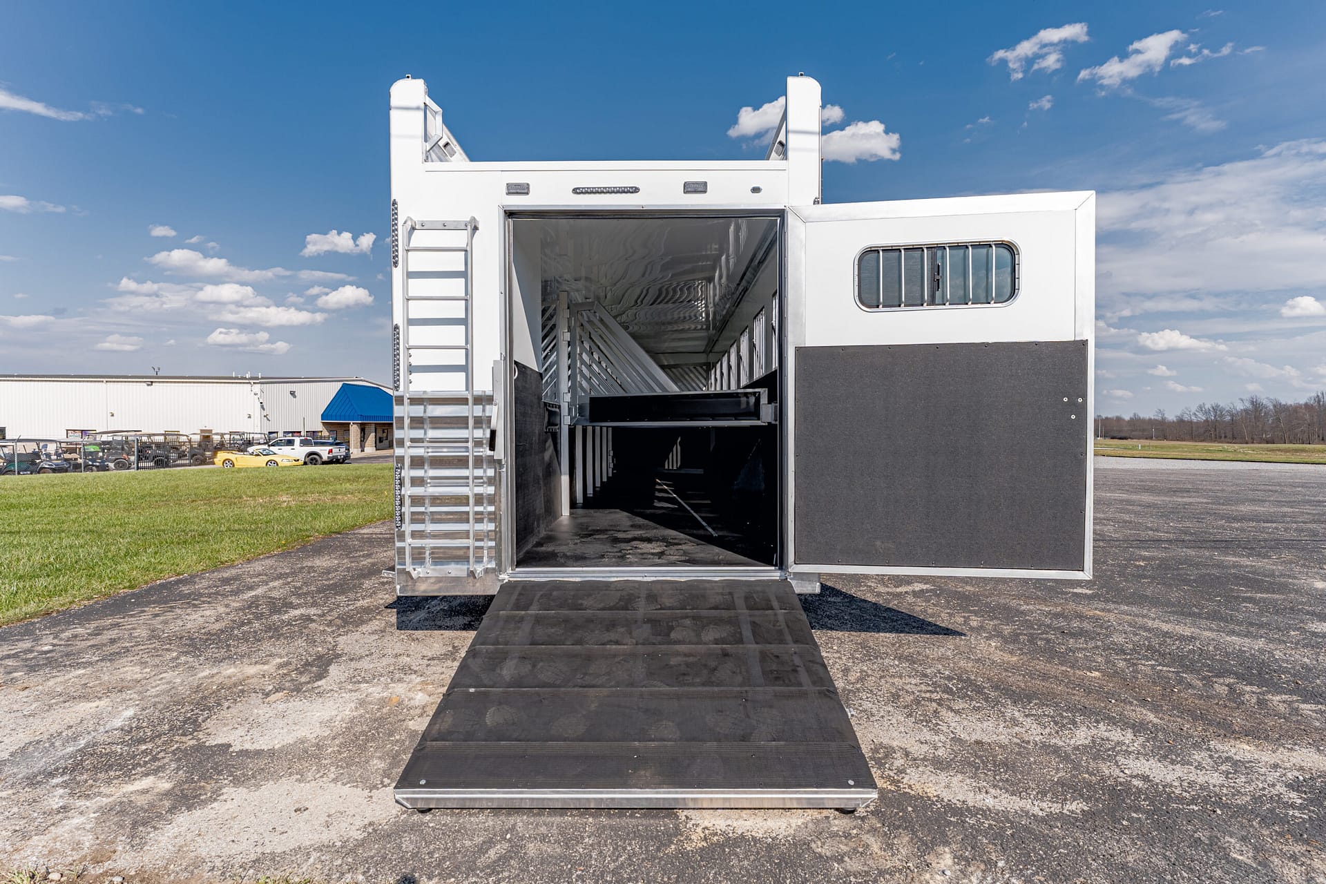 White livestock trailer with its rear doors open and a black loading ramp extended to the ground, showing interior stalls.