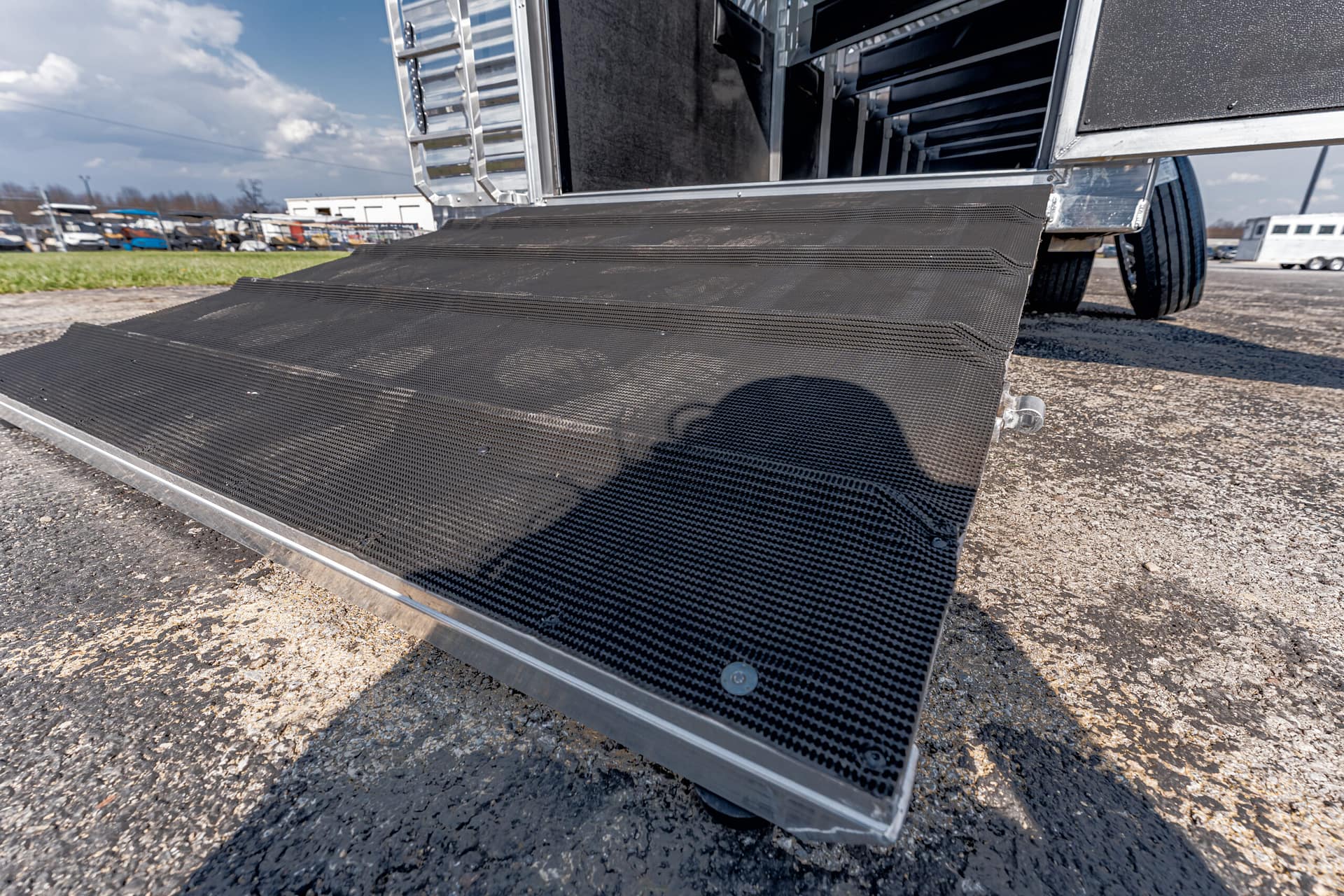 Black, non-slip loading ramp attached to a trailer, with a shadow of a person on its surface, outdoors on asphalt.