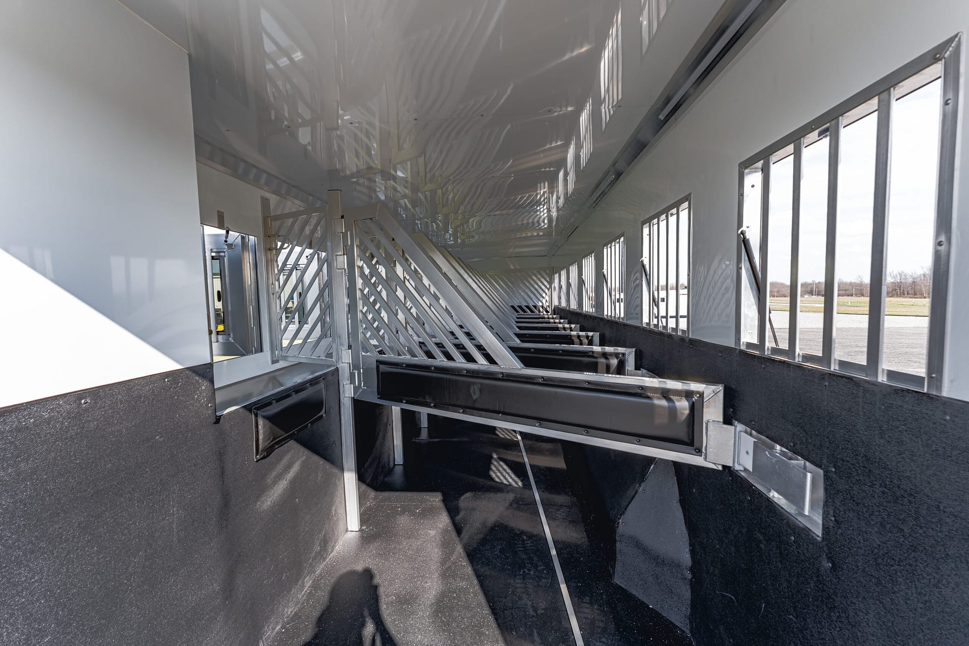 Interior of a metal trailer with slatted partitions and stalls along a black rubber floor, daylight streaming through side windows