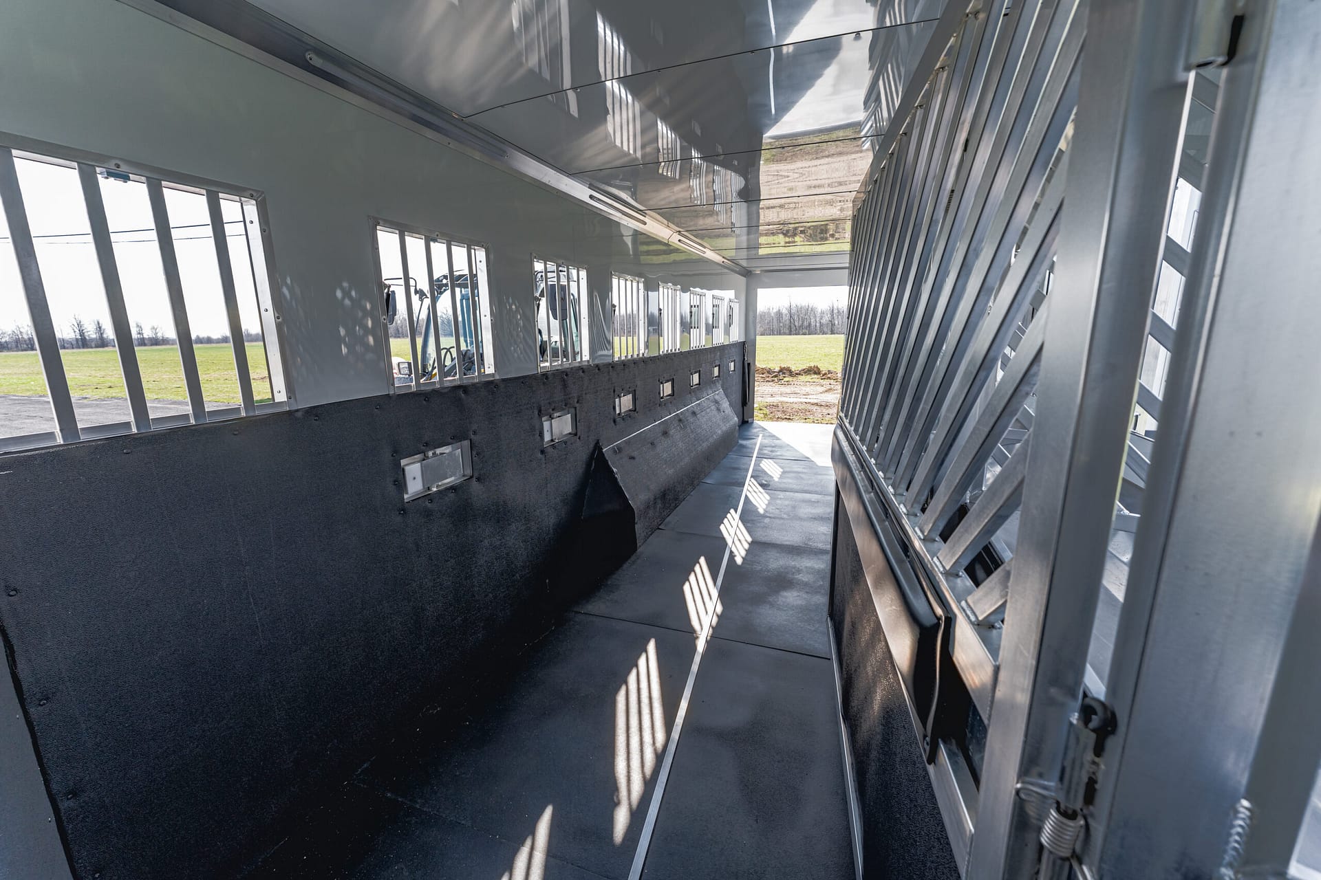 Interior of a metal livestock trailer with barred windows along the left side and a slatted divider on the right, sunlight from outside.