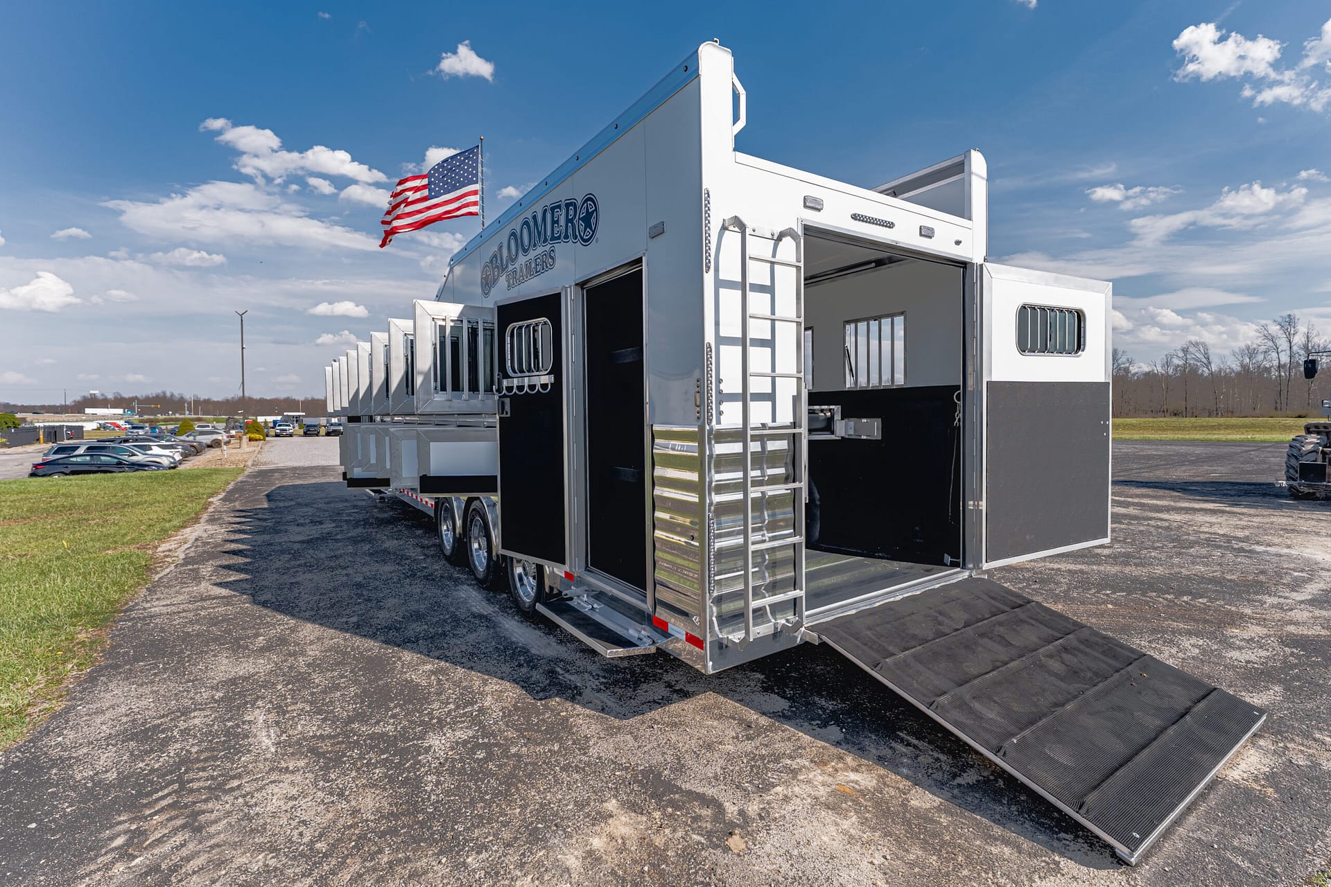 White multi-axle horse trailer with an open rear ramp and American flag flying above, photographed in a dealership lot.