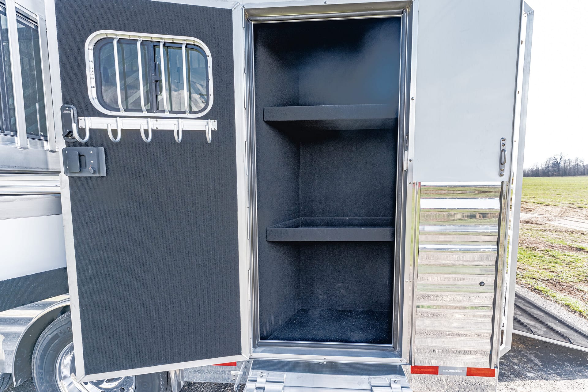 Open side storage compartment of a black-felt interior trailer with two shelves and a row of metal hooks on the door panel