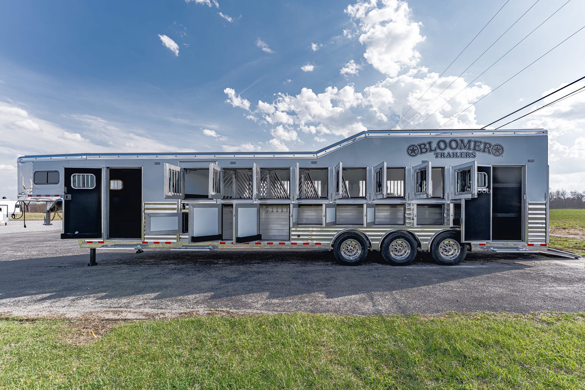 White horse trailer with numerous open stall doors and a Bloomers Trailers logo, parked on a gravel lot under a blue sky.