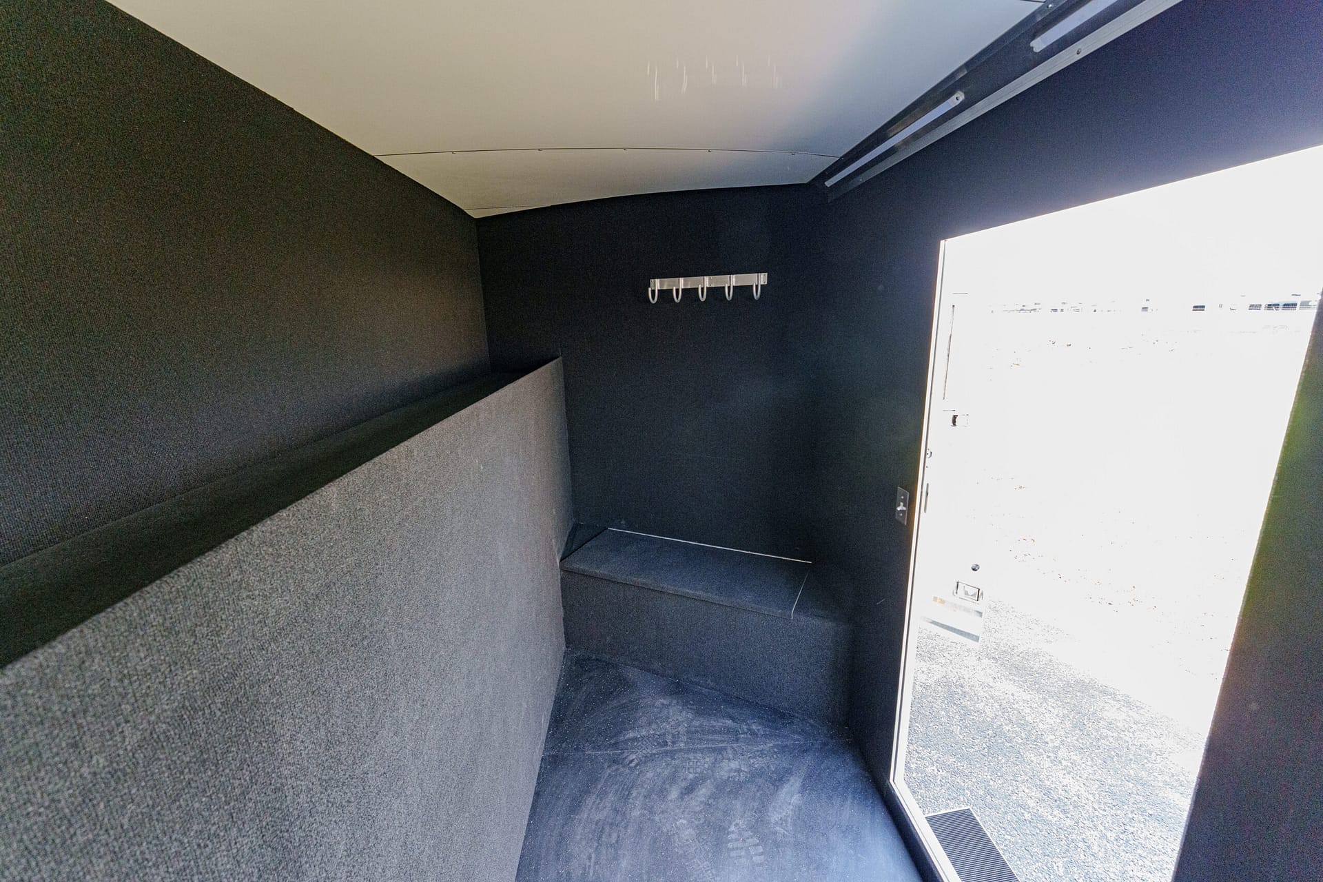 Dark, narrow interior with a padded bench along the back wall and a row of metal coat hooks, doorway open to the outside on the right.