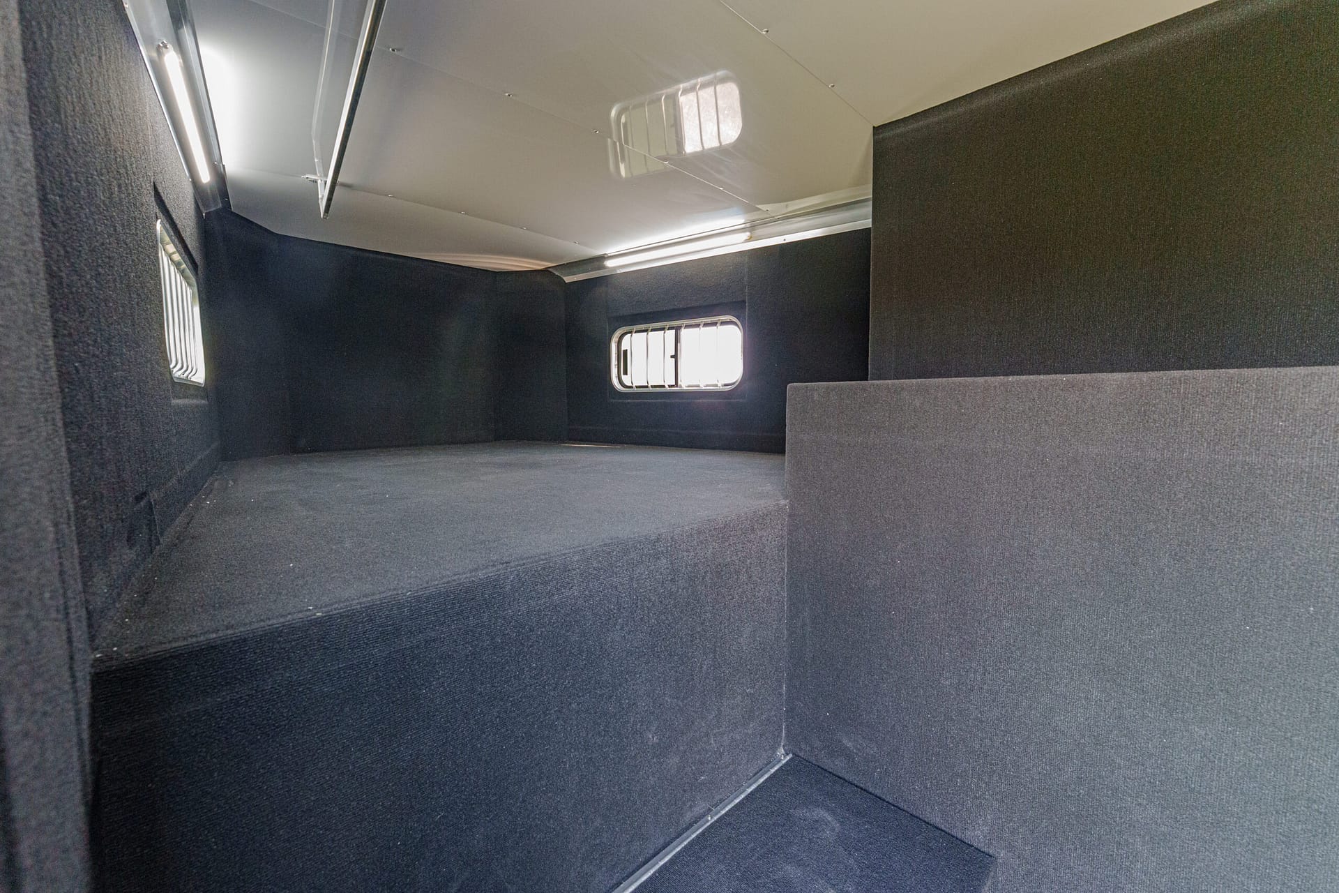 Dark cargo trailer interior with black fabric walls, a raised platform along the left, and small rectangular windows with bars on the walls
