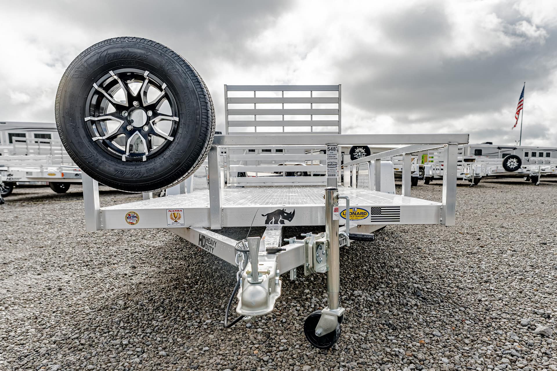 Aluminum utility trailer with a mounted spare tire on the front, parked on a gravel lot among other trailers far in the background.