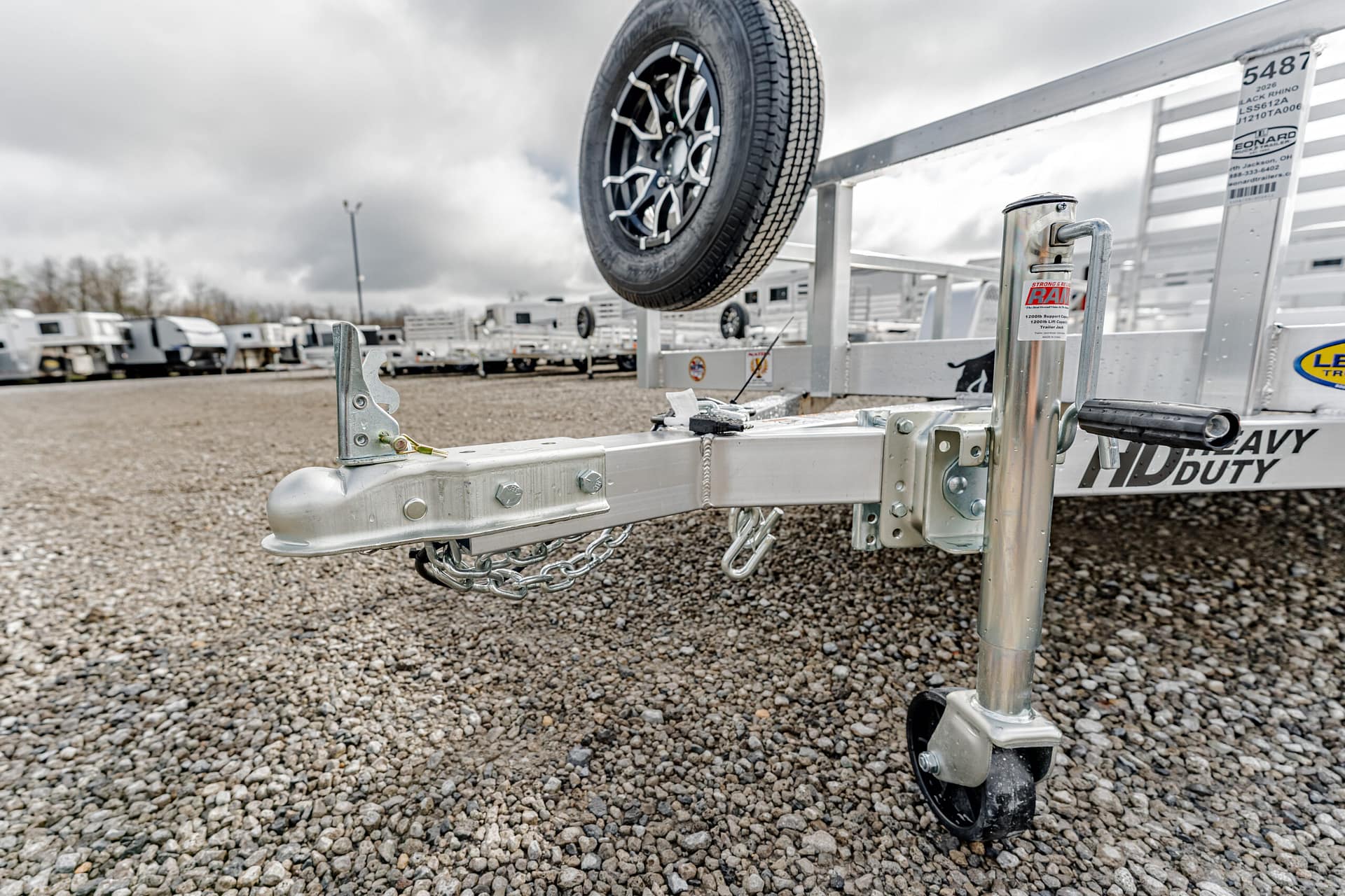 Close-up of a trailer hitch and safety chains on a metal utility trailer, with a spare tire mounted overhead on a gravel lot.