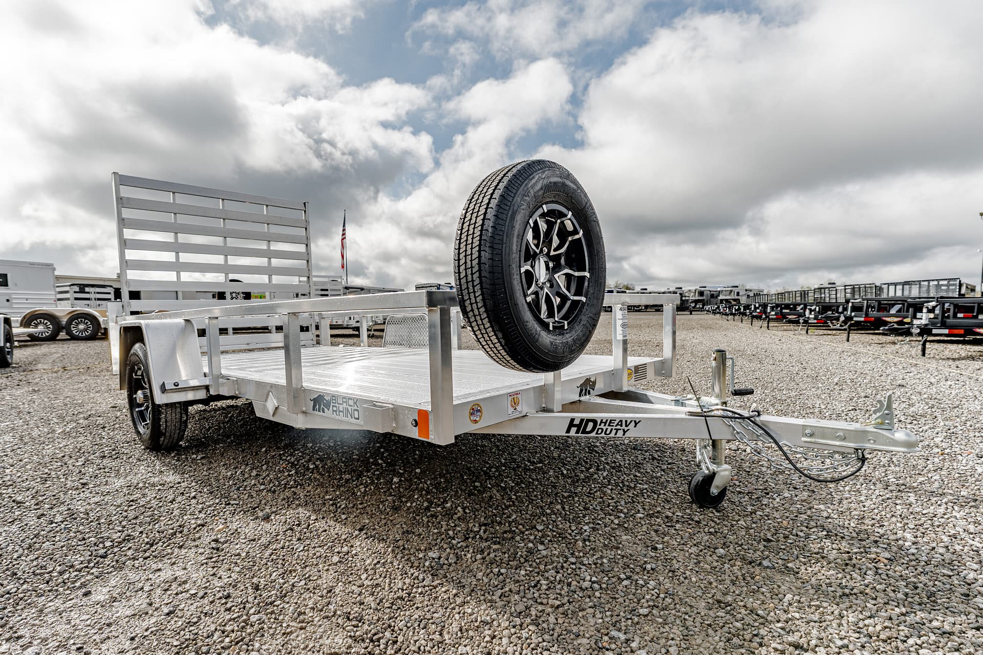 Aluminum flatbed utility trailer with a mounted spare tire on the front, parked on a gravel lot.