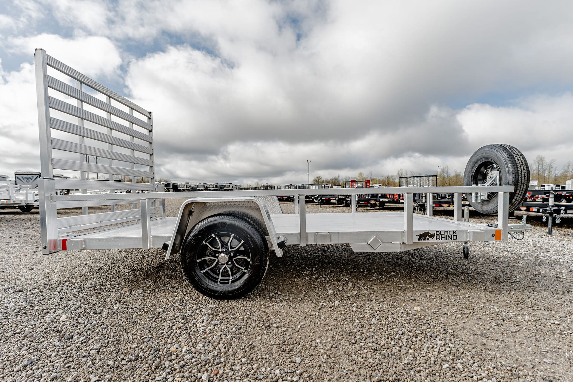 Aluminum flatbed trailer with tall ramp gate on a gravel lot, spare tire mounted at the front right, Black Rhino logo