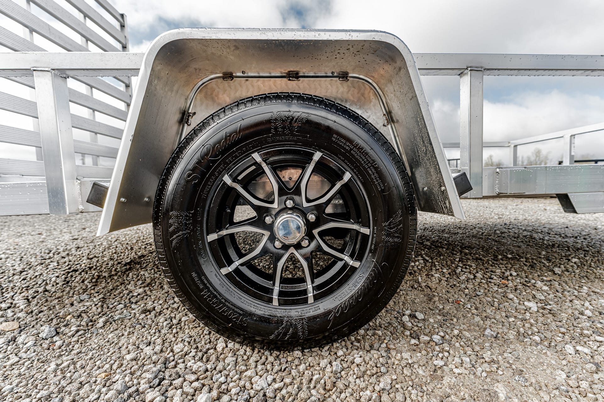 Close-up of a black alloy trailer wheel with chrome center cap and metal fender on a gravel lot/grounded in a trailer setup.