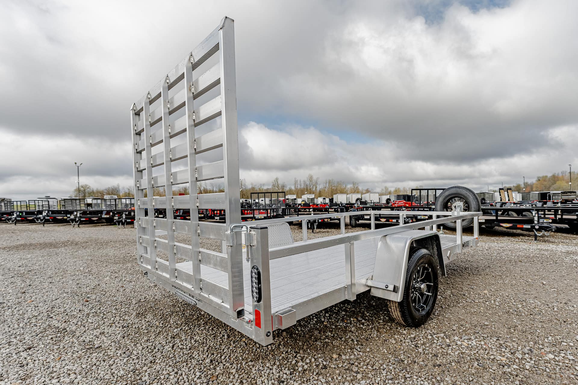 Silver aluminum trailer with a tall, vertical gate on a gravel lot, other trailers in the background.