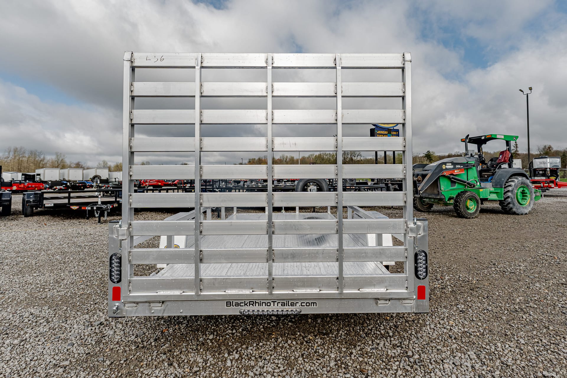 Rear view of a silver metal utility trailer with a slatted gate and tail lights, parked on gravel; 'BlackRhinoTrailer.com' visible on bumper.