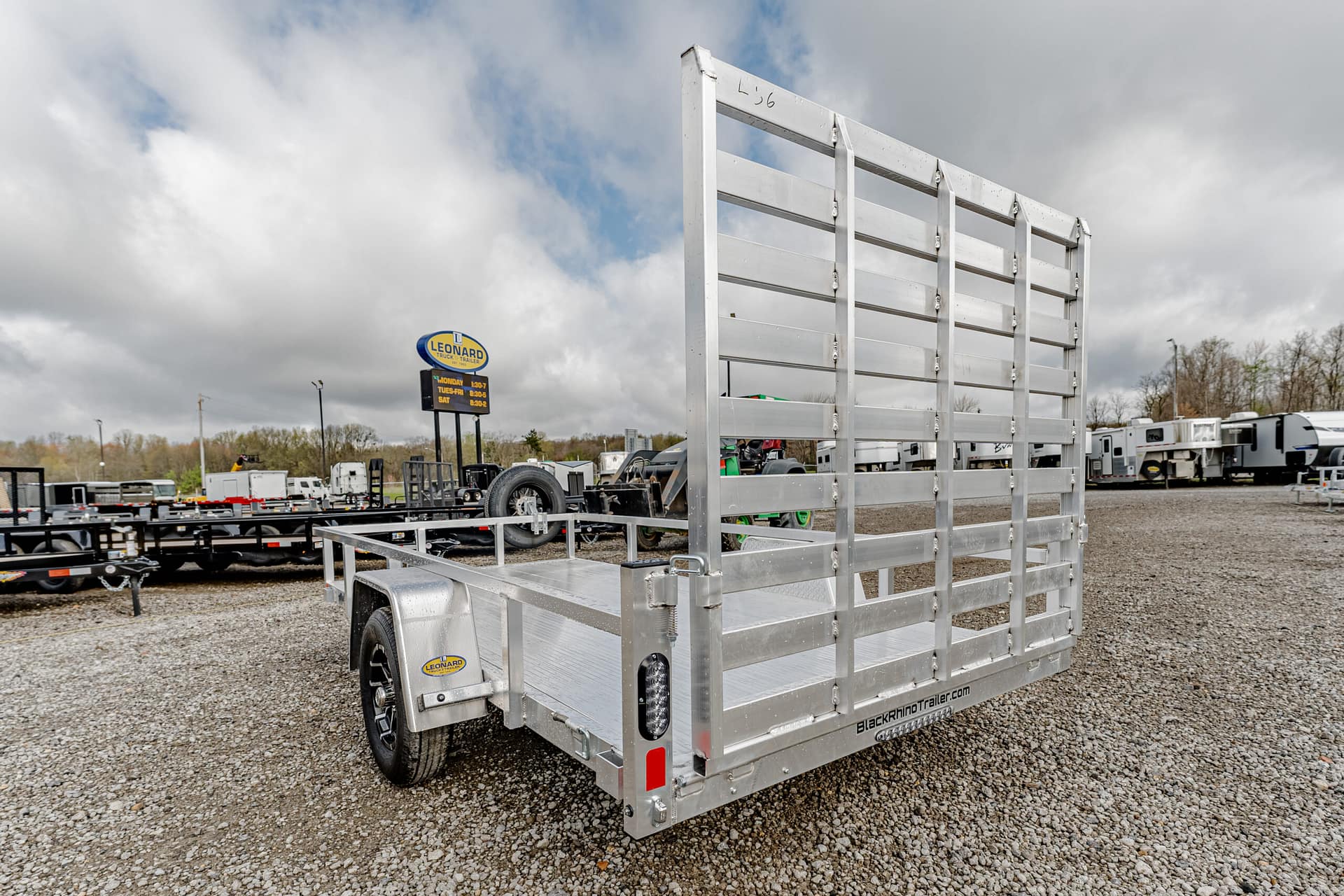 Aluminum utility trailer with a tall gate on a gravel lot, surrounded by other trailers and RVs.