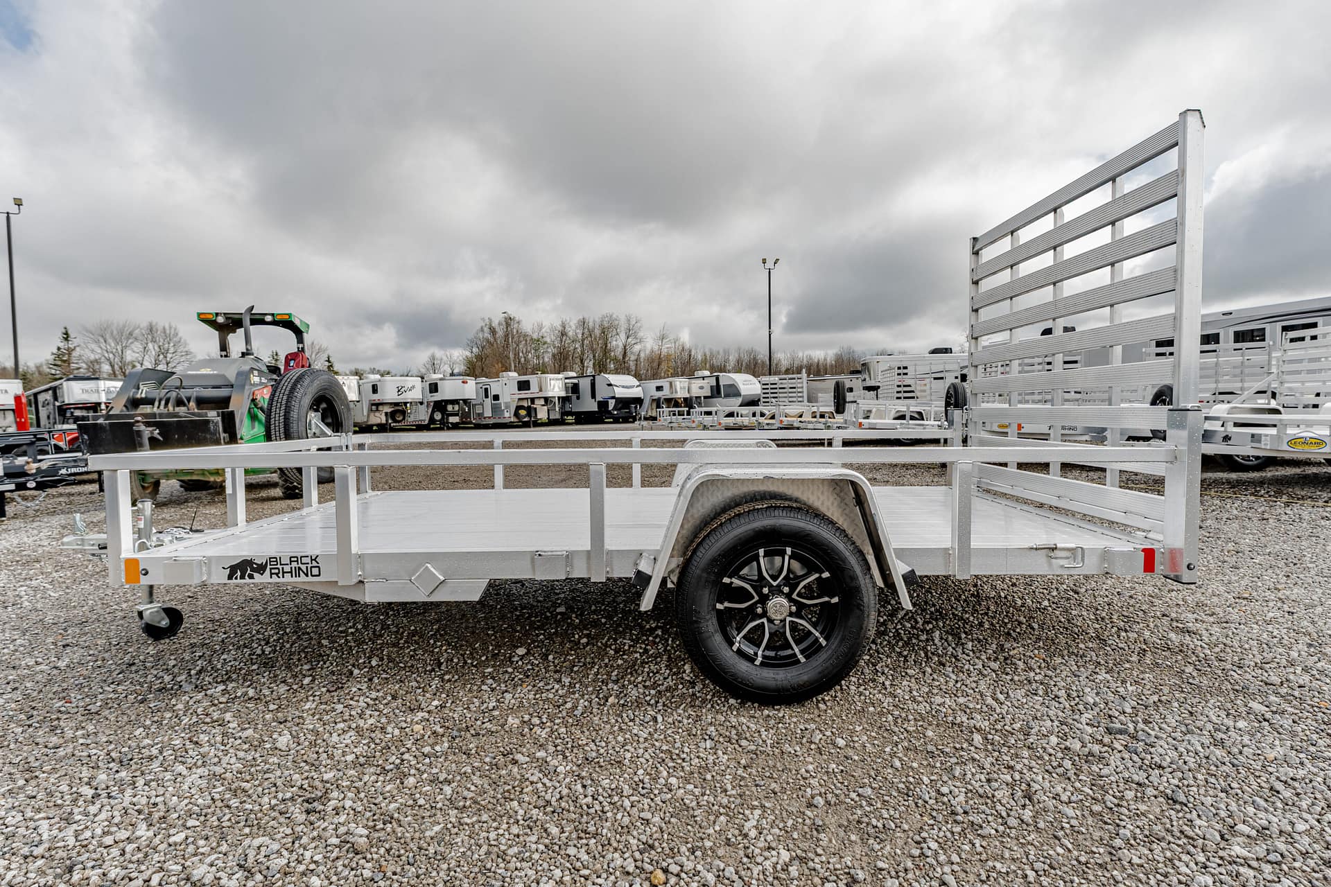 Silver aluminum flatbed trailer with a tall ramp gate, parked on a gravel lot among other trailers.