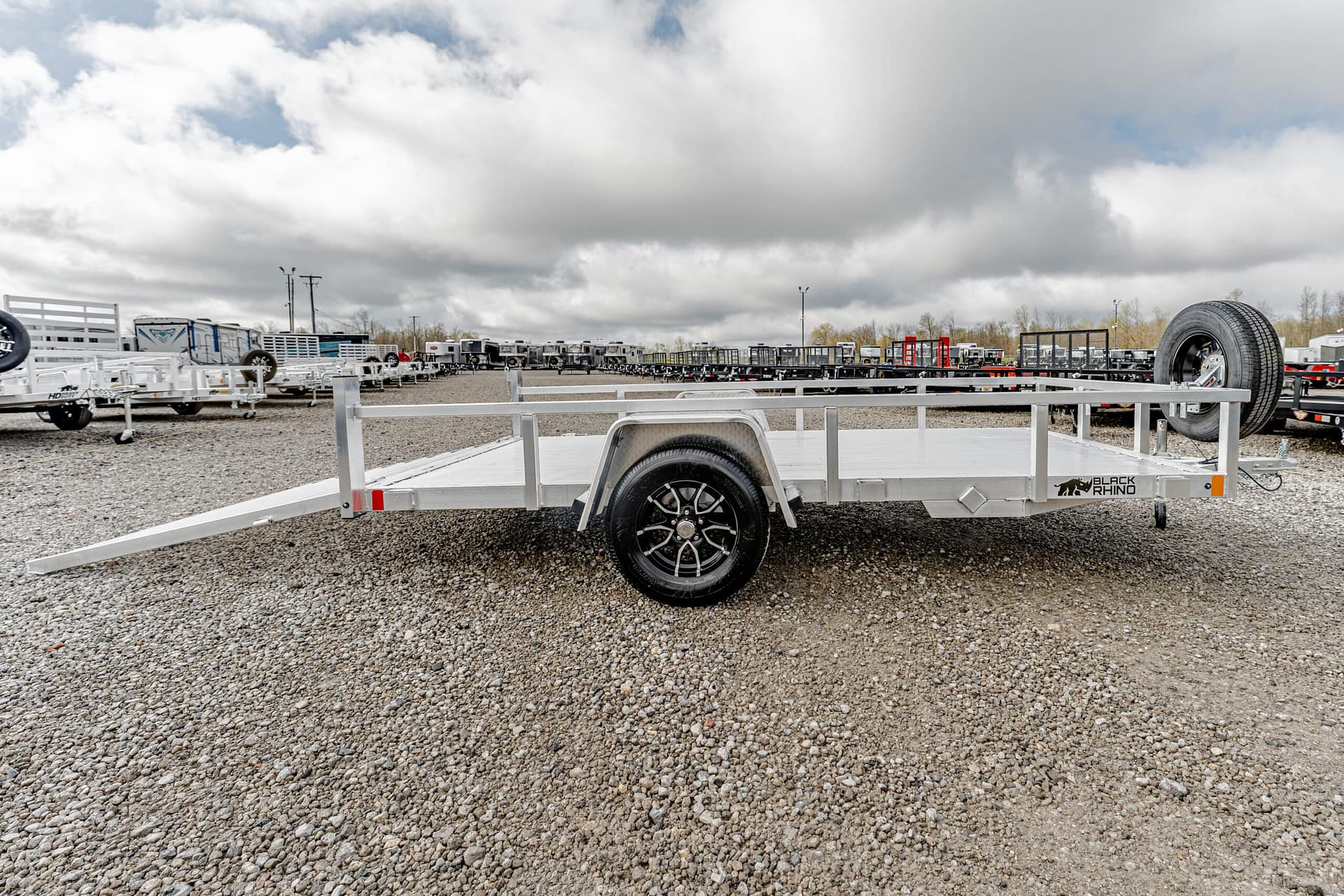 Aluminum flatbed trailer with side rails on a gravel lot, spare tire mounted at the rear and a Black Rhino logo visible on the side.