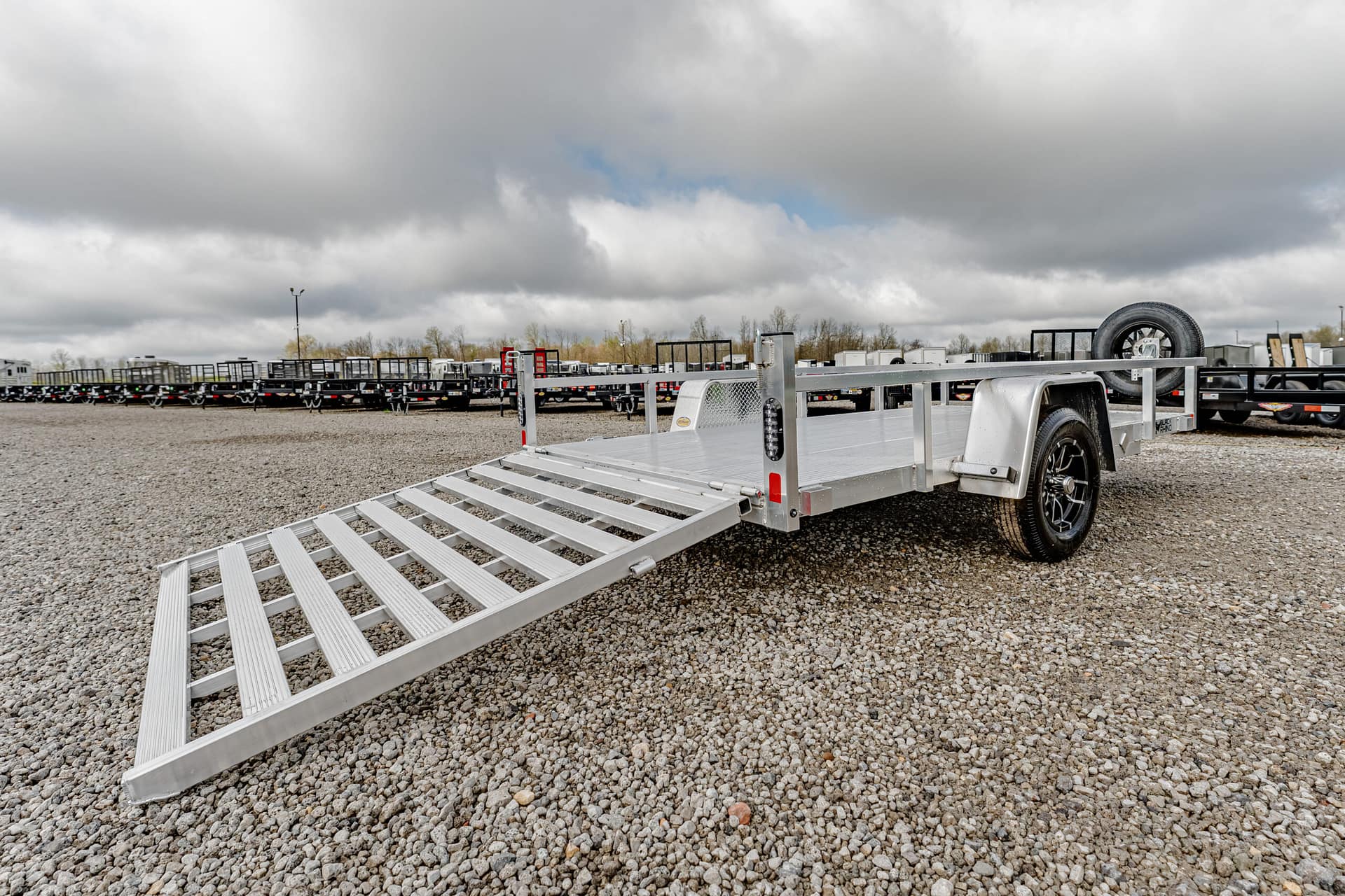 Silver aluminum utility trailer with a lowered ramp on a gravel lot, other trailers visible in the background under a cloudy sky.