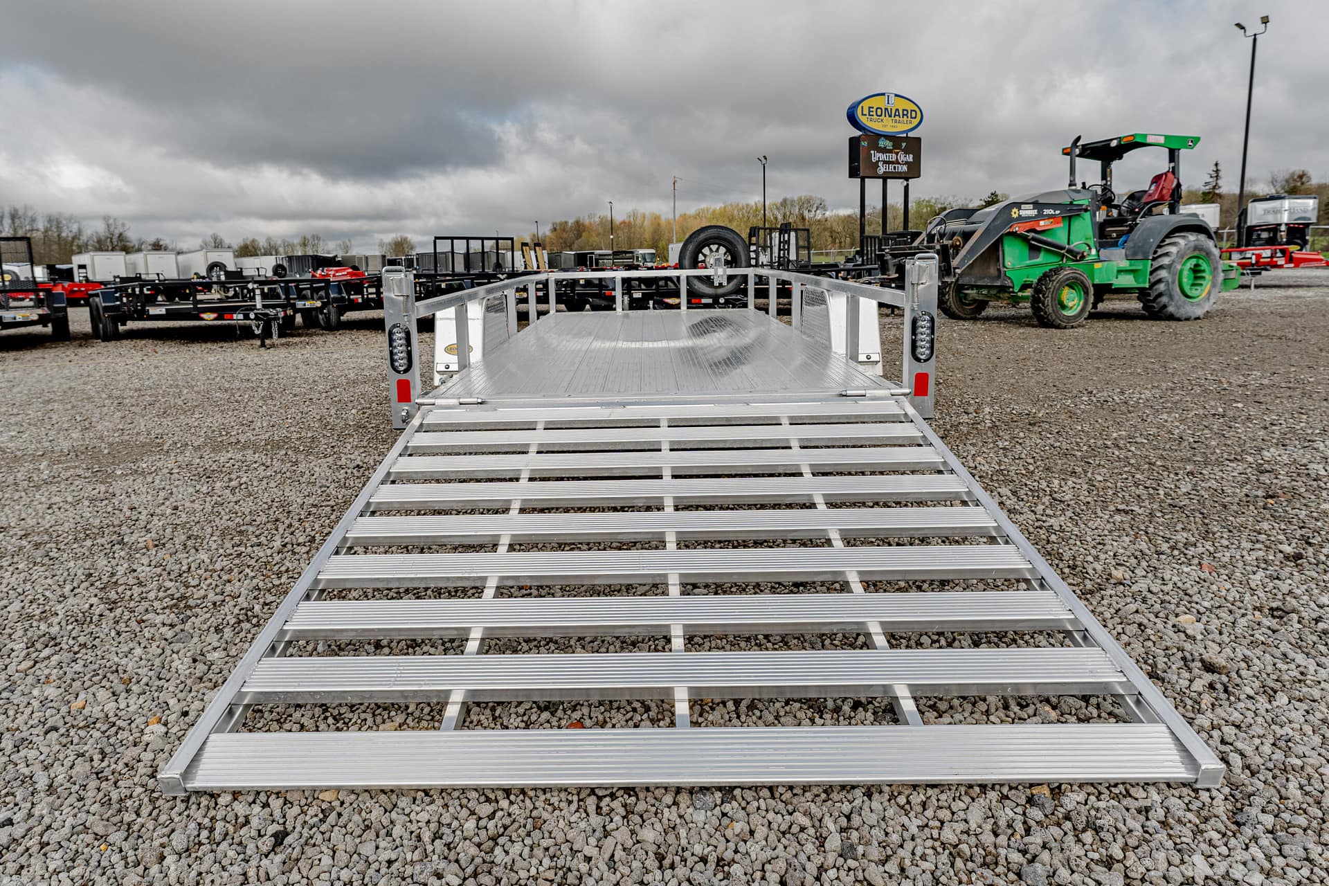 Aluminum equipment trailer with a lowered ramp on a gravel lot, ready for loading.