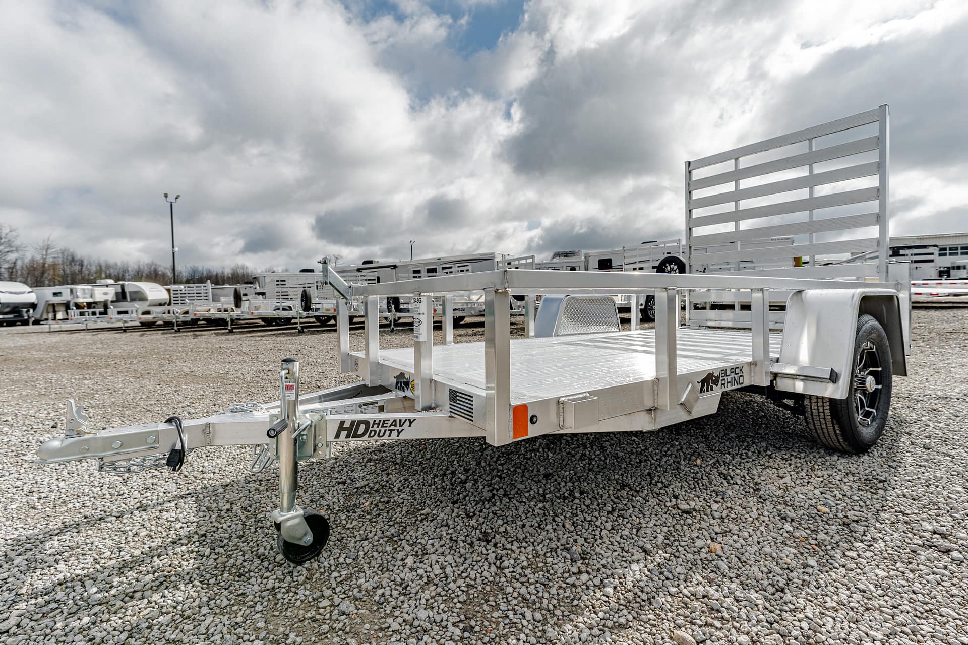 Aluminum flatbed trailer on a gravel lot with other trailers in the background and cloudy sky above.