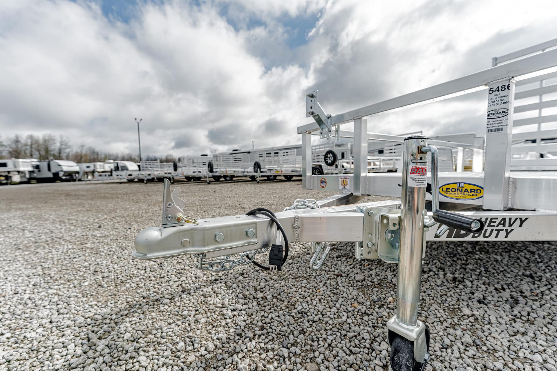 Close-up of a metal trailer hitch and stabilizing jack on a gravel lot, with rows of white trailers in the background.