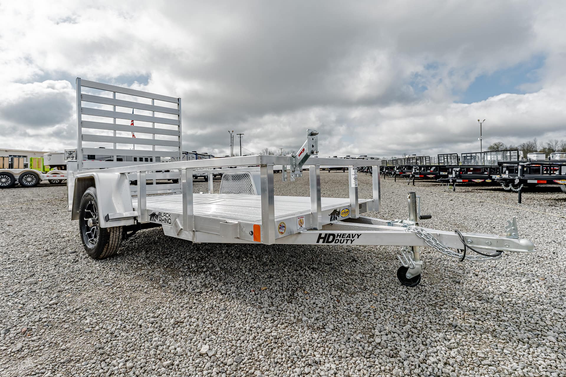 Silver heavy-duty flatbed trailer on a gravel lot with other trailers in the background under a cloudy sky.