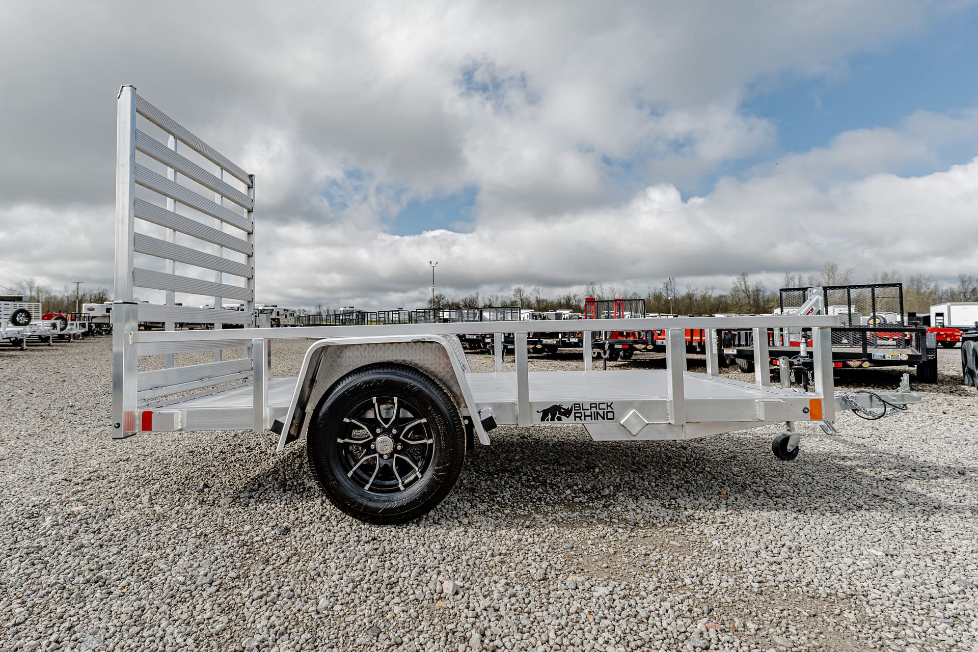 Aluminum utility trailer with a tall ladder rack on a gravel lot, other trailers in the background under a cloudy sky