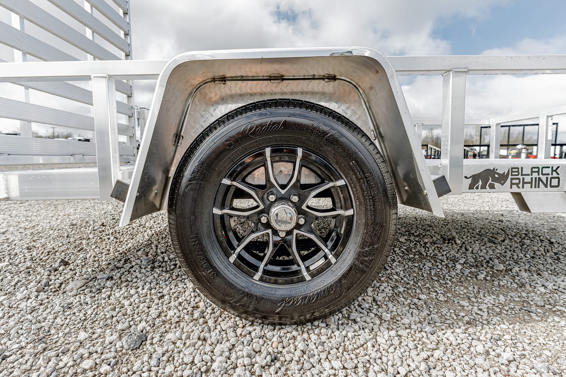 Close-up of a black alloy trailer wheel inside a curved metal fender, with a gravel lot and a white trailer frame in the background; a Black Rhino logo is visible on the side.