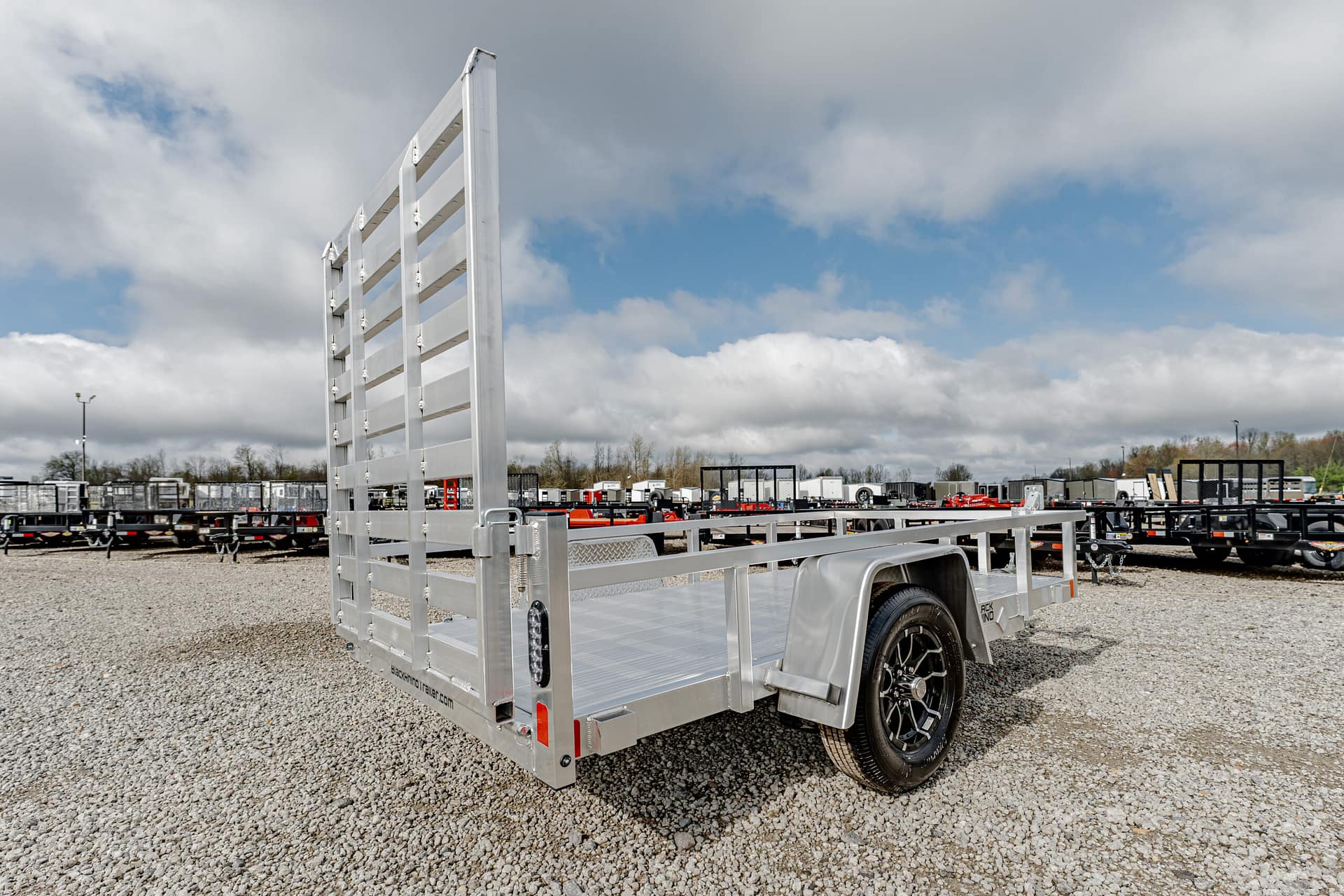Silver aluminum utility trailer with a tall ramp gate on a gravel lot, other trailers visible in the background under a cloudy sky.
