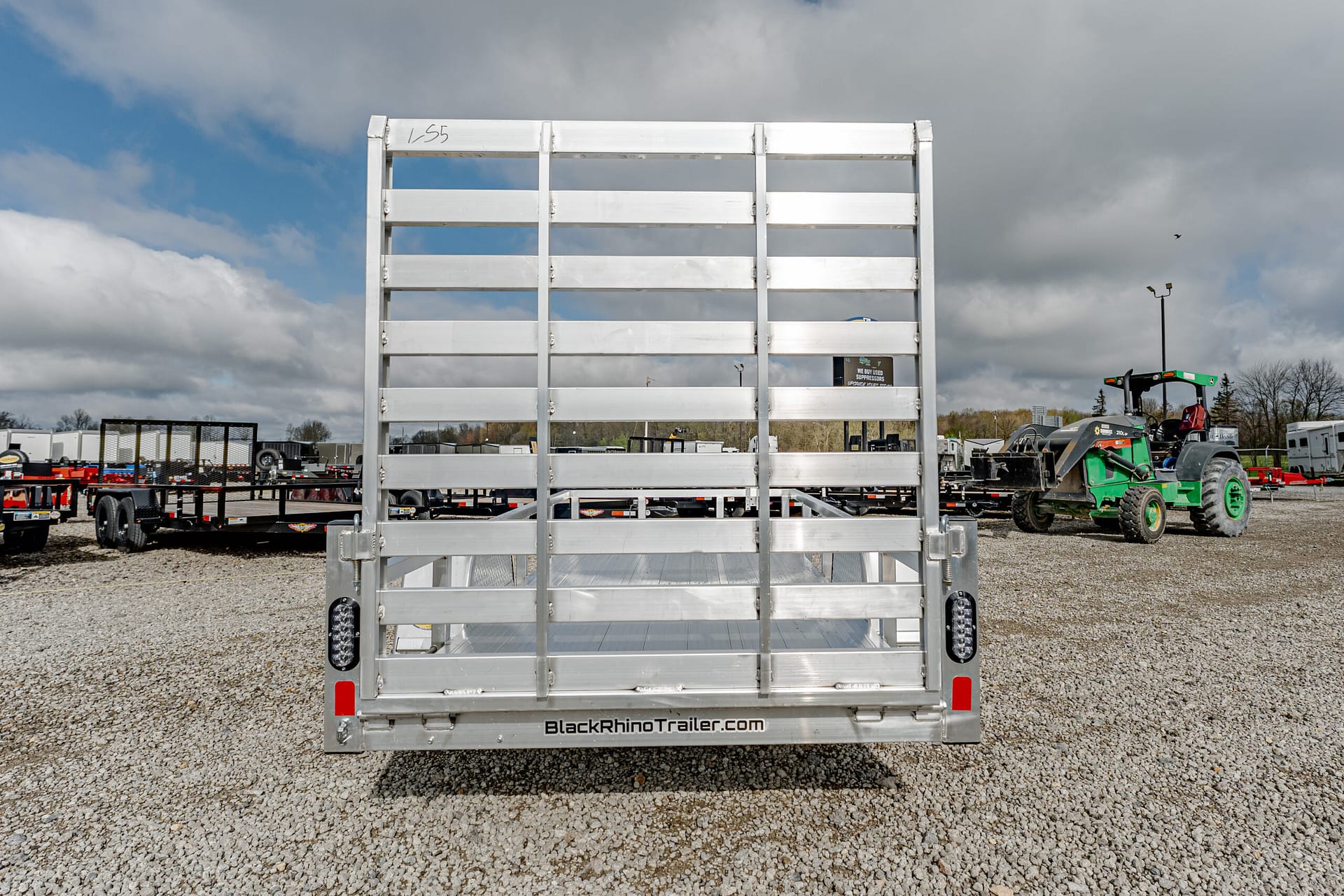Rear view of a silver aluminum trailer gate on a gravel lot, with machinery and a tractor in the background, under a cloudy sky.