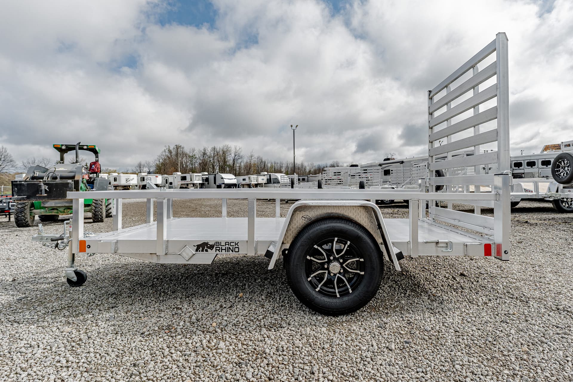 Flatbed utility trailer with tall side rails in a gravel lot, Black Rhino branding visible on the side wheel area.