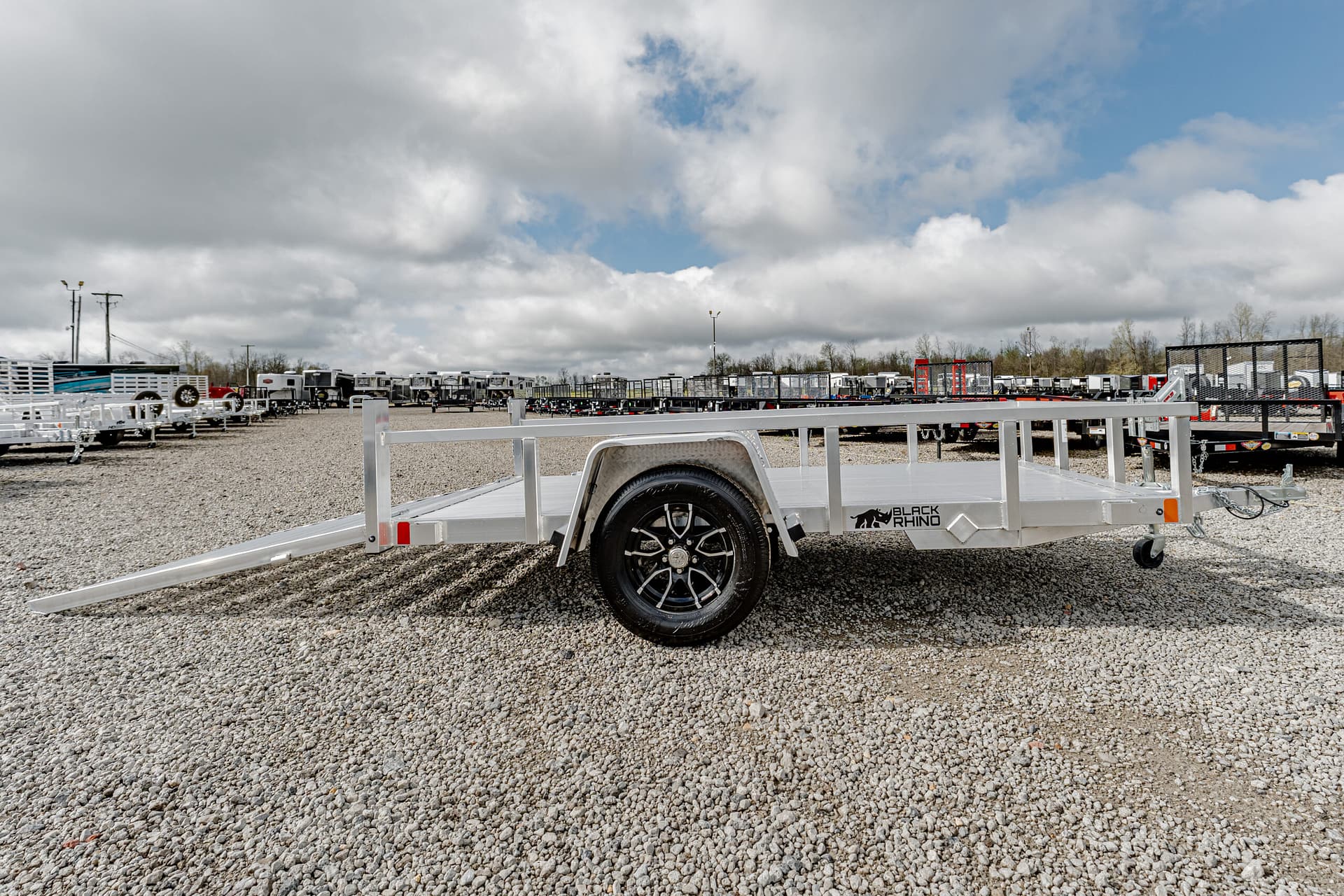 Row of aluminum flatbed trailers parked on a gravel lot beneath a cloudy sky, foreground shows a trailer with a Black Rhino logo.