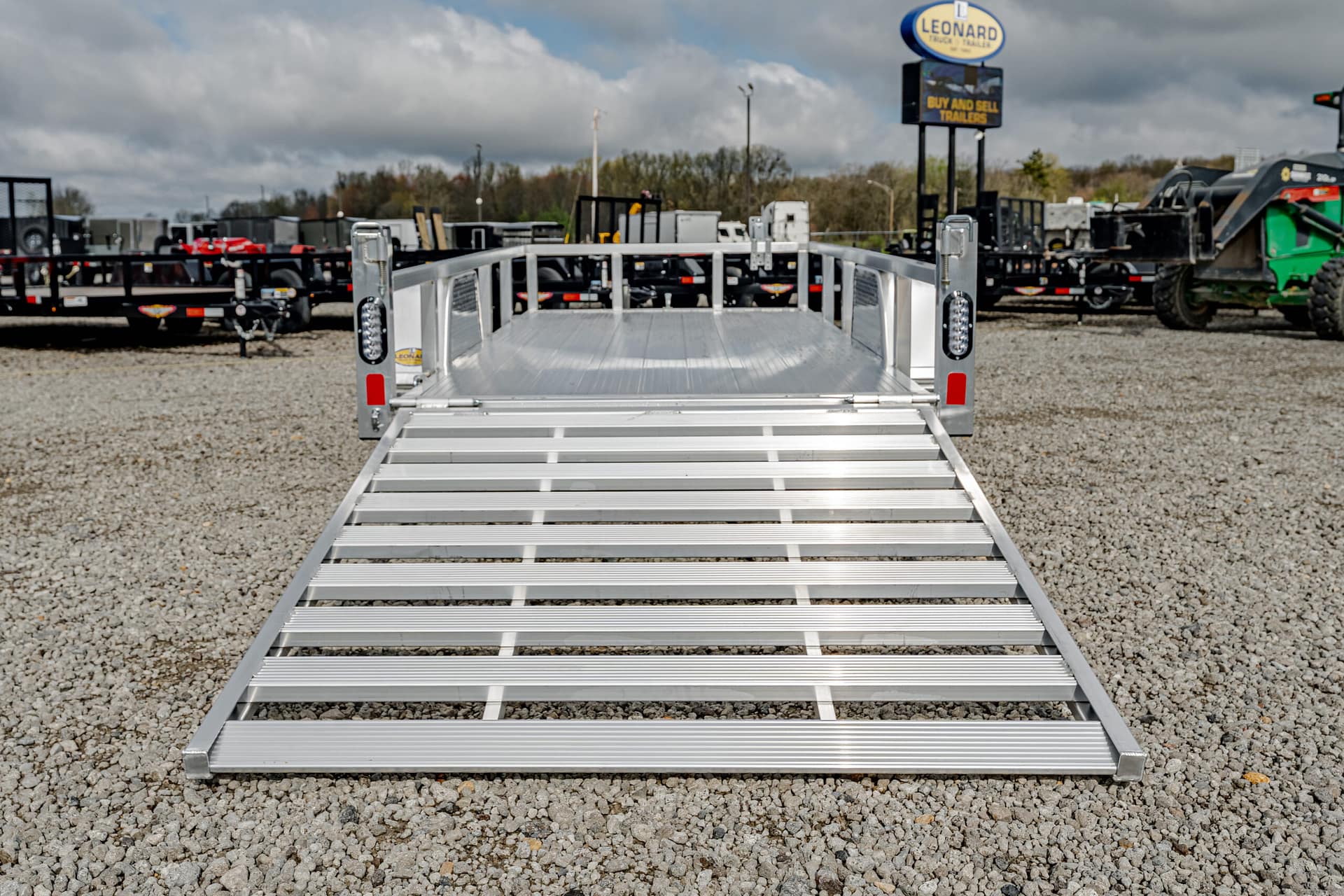 Aluminum trailer ramp extends toward the camera in a gravel lot with other trailers and equipment in the background.