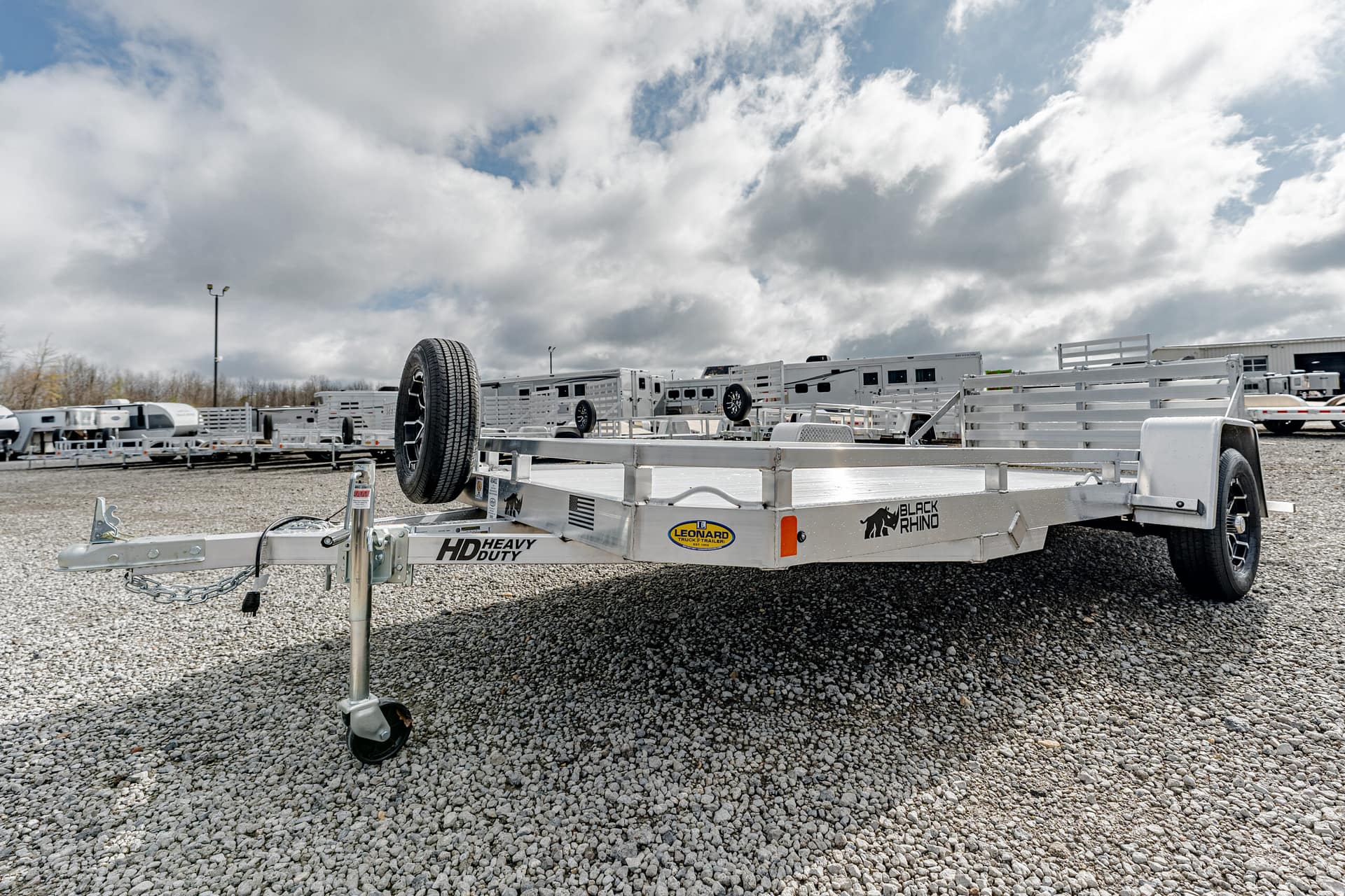 Aluminum flatbed trailer with a spare tire mounted on the front, parked on a gravel lot among other trailers.