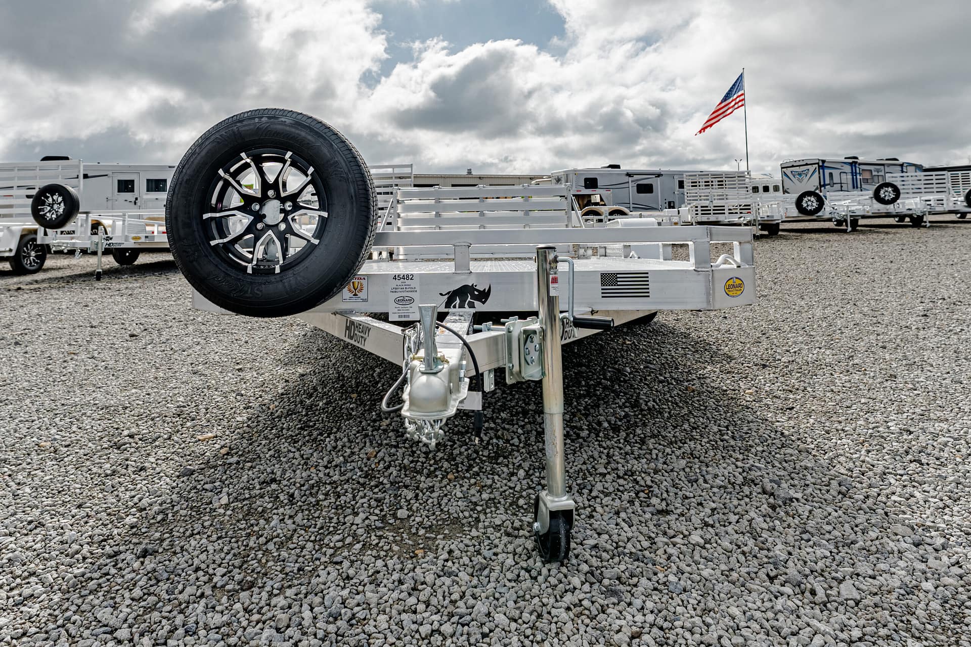 Front view of a silver trailer with a mounted spare tire on a gravel lot, RVs in the background and an American flag in the distance.