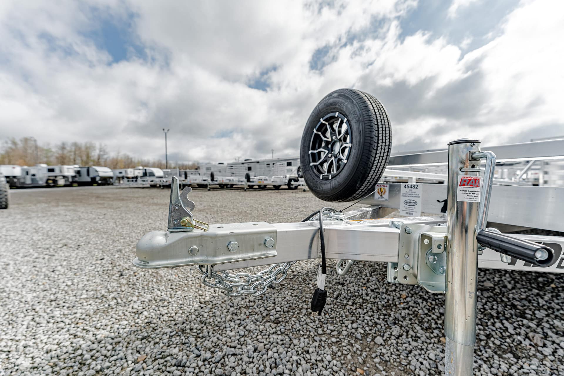 Close-up of a silver trailer tongue with a mounted spare tire and safety chains on a gravel RV lot.23%